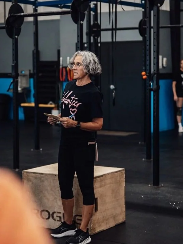 A woman with gray curly hair and glasses, wearing a black T-shirt with 'Hunter' written on it and black leggings, stands in a gym near a wooden box, holding a notepad and pen, with gym equipment in the background.