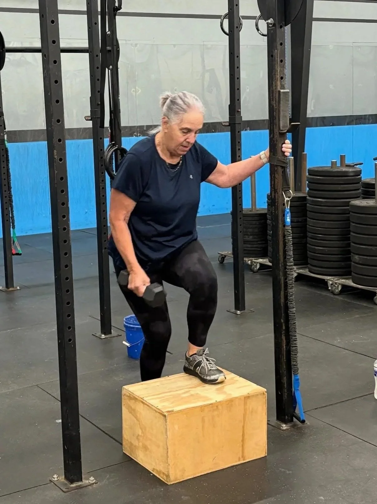A woman standing on a wooden box lifting a dumbbell in a gym.