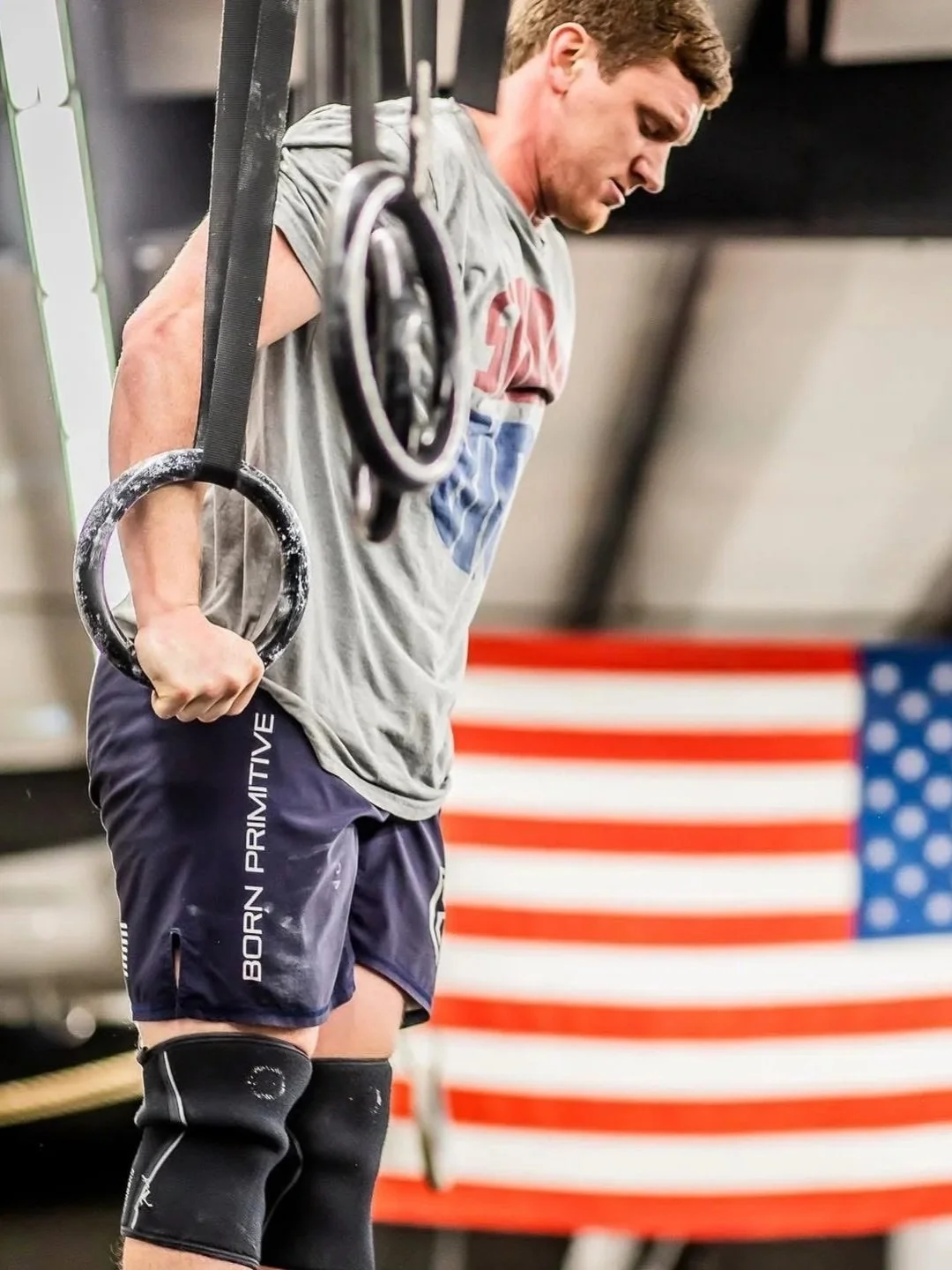 A man in athletic attire performing a dip exercise on gym rings, with an American flag in the background.