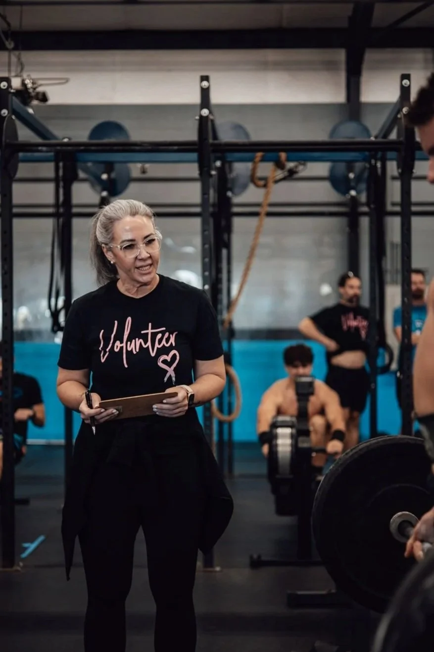 A woman wearing a black shirt with the word 'Volunteer' and a heart graphic is standing in a gym, holding a clipboard, with a barbell in front of her and exercise equipment and people in the background.