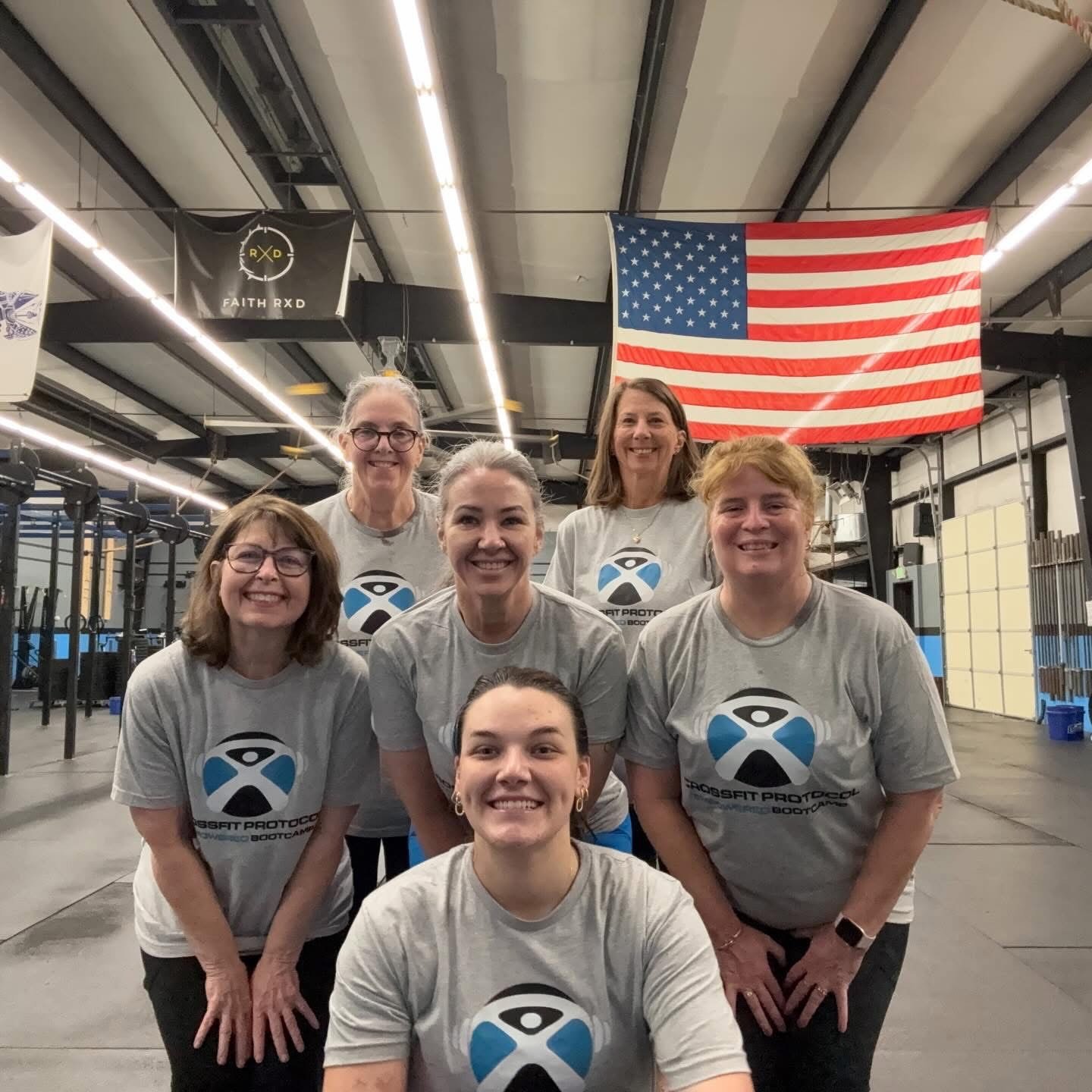 Six women posing together in a gym, wearing matching gray t-shirts with a logo and text that reads "CrossFit Protocol Bootcamp." An American flag hangs on the wall behind them, and gym equipment can be seen in the background.
