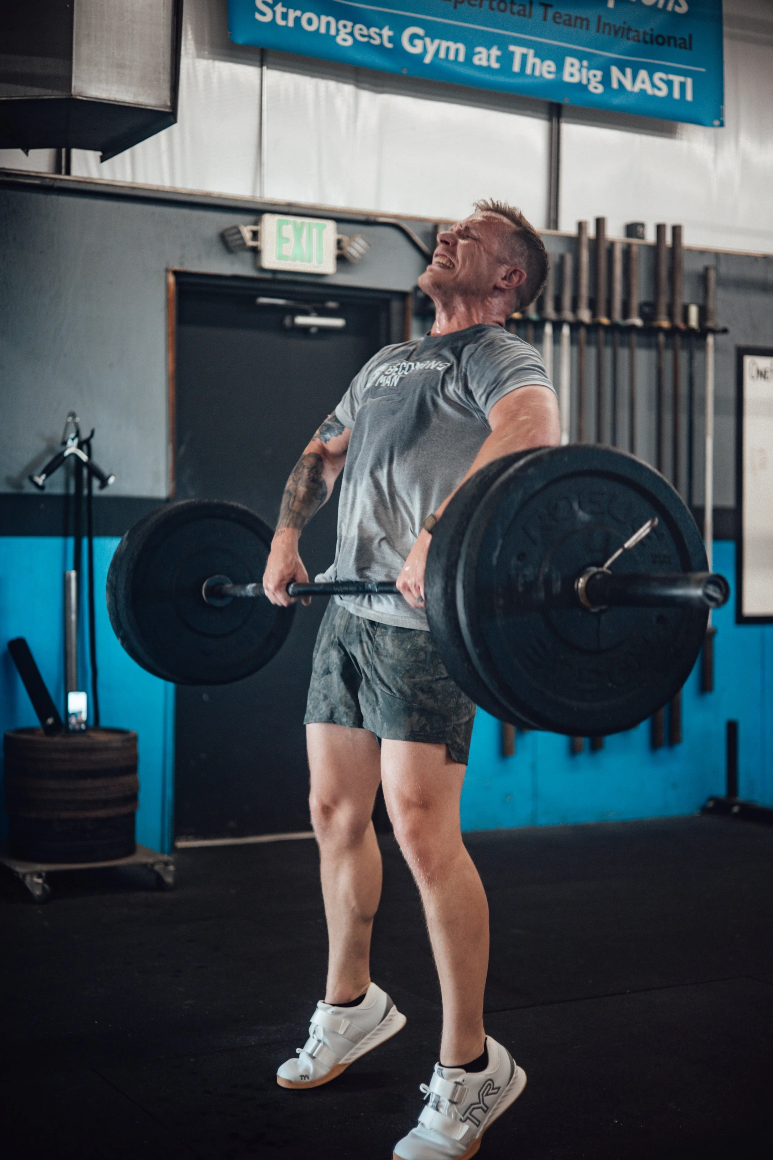 Man lifting a heavy barbell with weights at a gym, showing effort. Gym environment with blue wall, hanging weights, and a blue banner overhead.