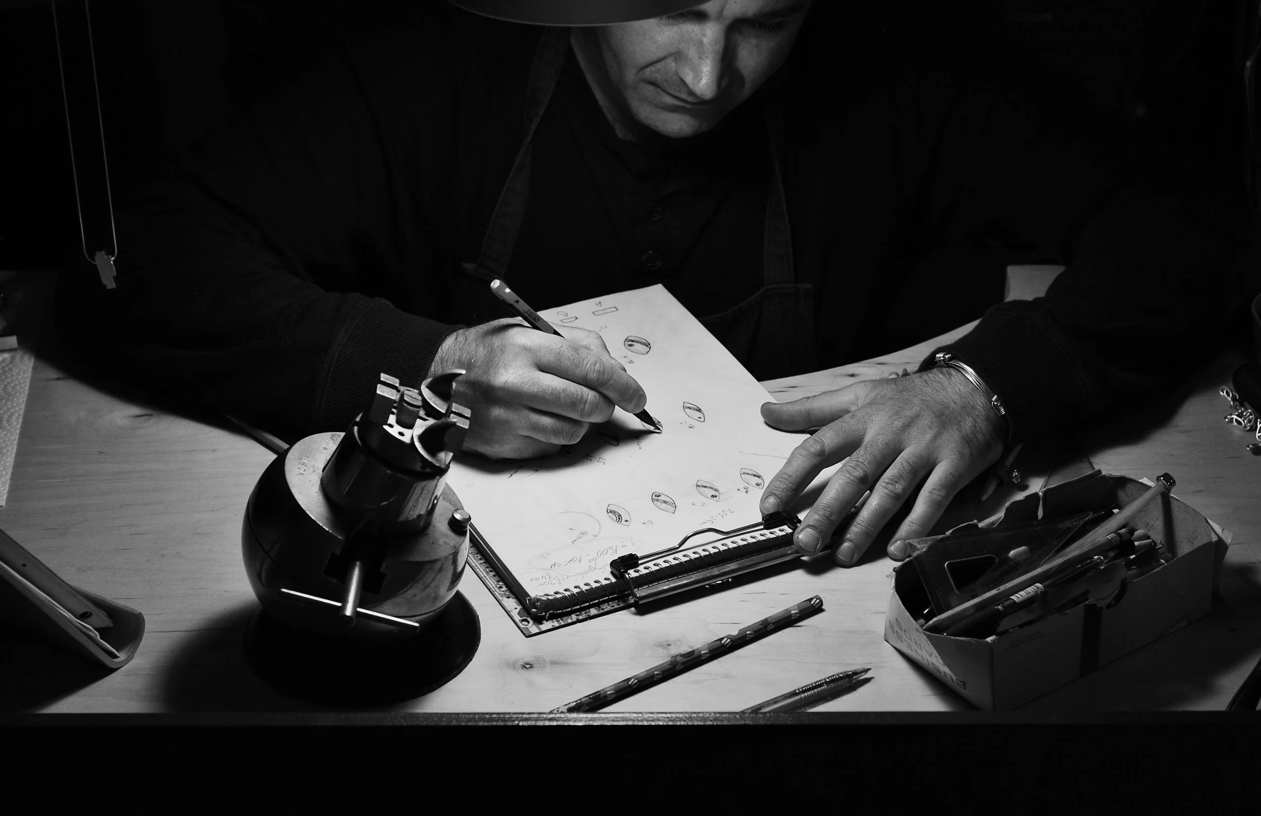 Zsolt Szekely sketches on a sheet of paper, surrounded by various tools and objects including a microscope, pens, and electronic components.