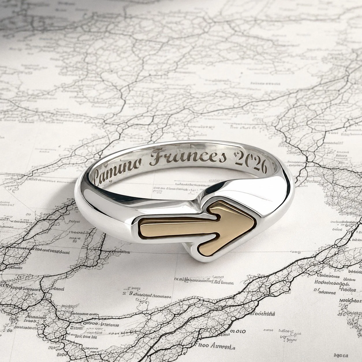 Silver ring engraved with "Camino Frunces 2026" and an arrow symbol, placed on a map with dark lines depicting a network of paths or routes.