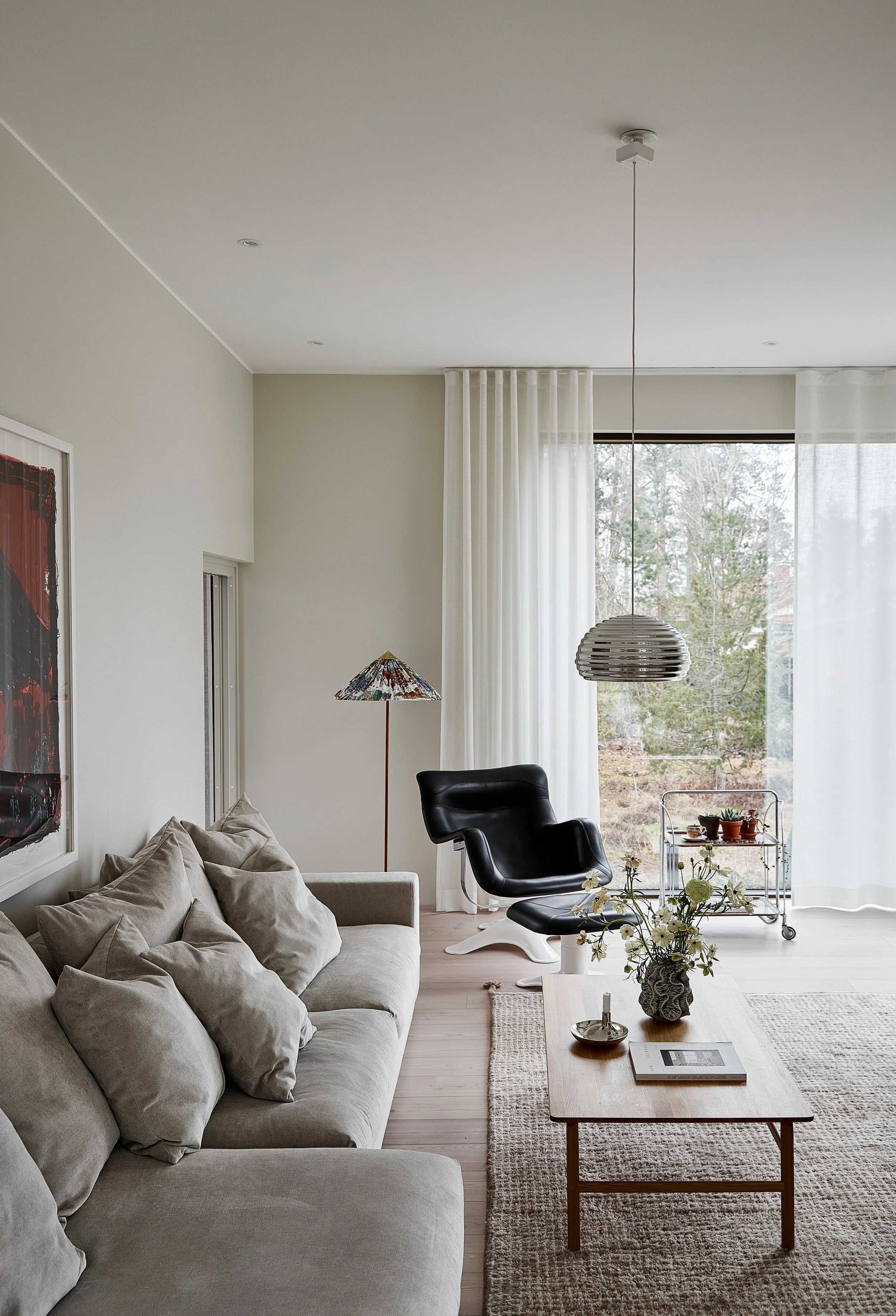Living room with beige sofa, black lounge chair, wooden coffee table, floor lamp, pendant light, and large window with white curtains.