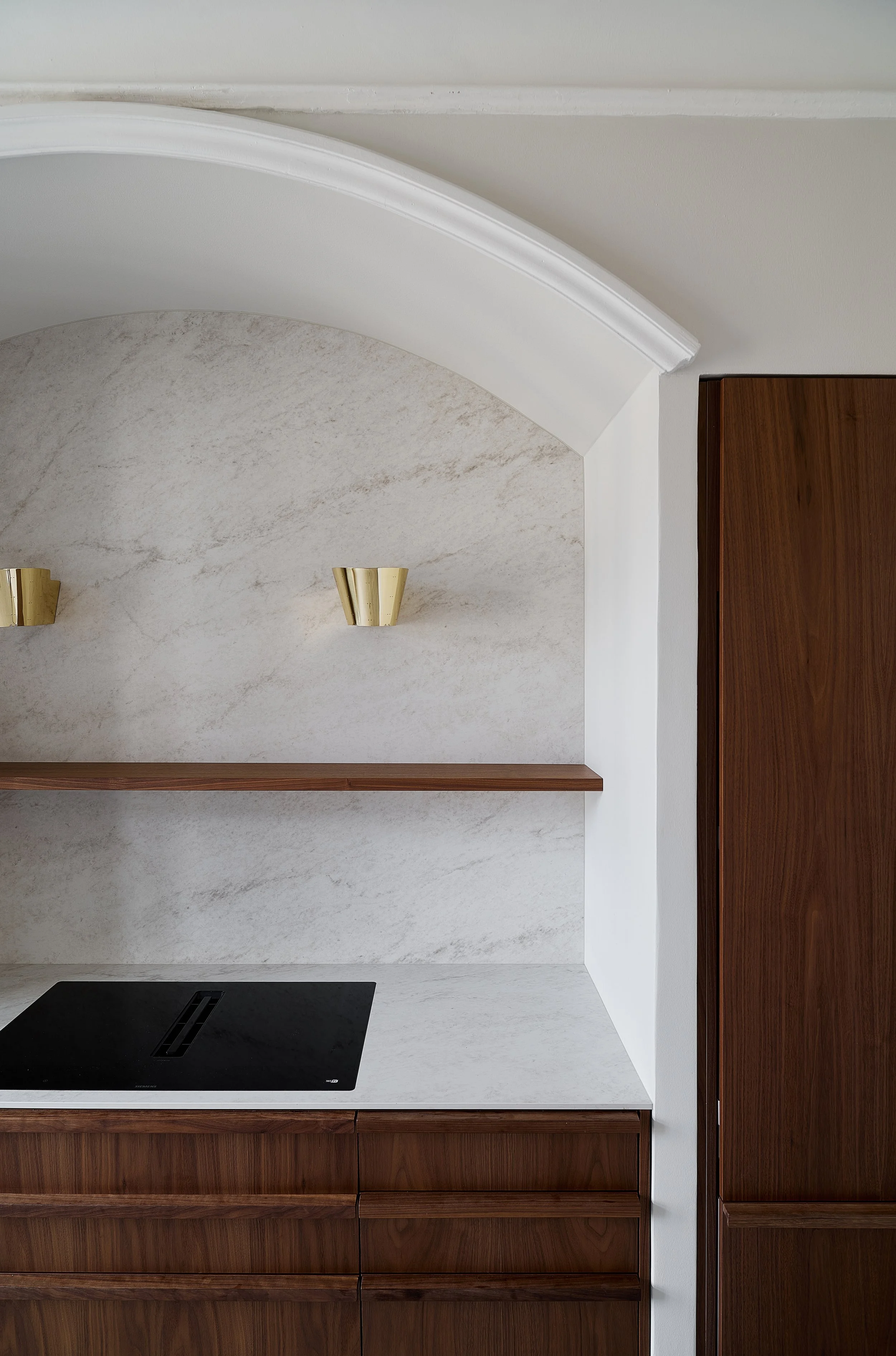 A minimalist custom kitchen interior with marble backsplash, dark wood cabinets, a black electric cooktop on a white marble countertop, and two gold wall sconces.