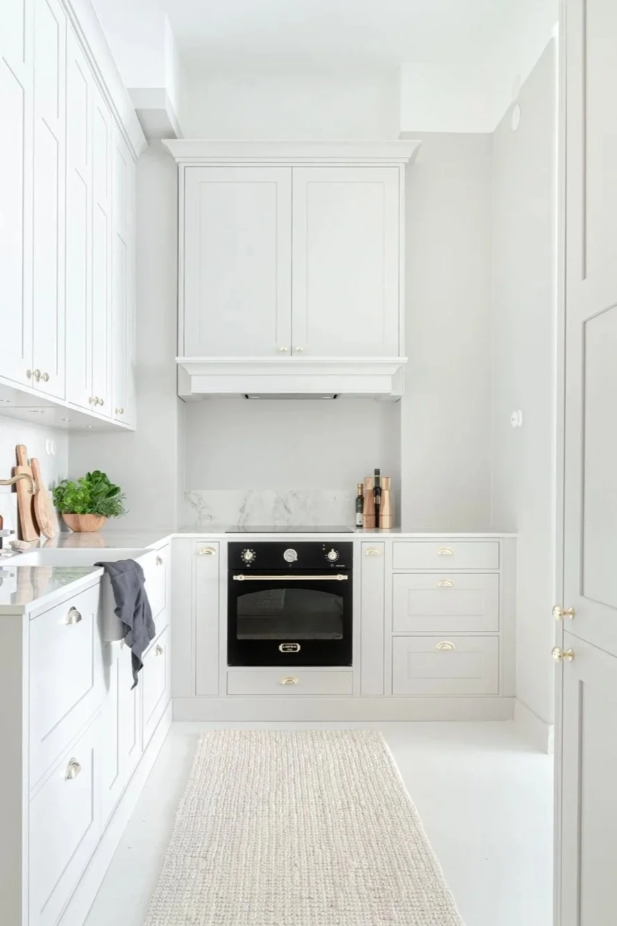 Bright, white kitchen with built-in cabinets, a black oven, marble backsplash, green potted plant, and wooden kitchen utensils.