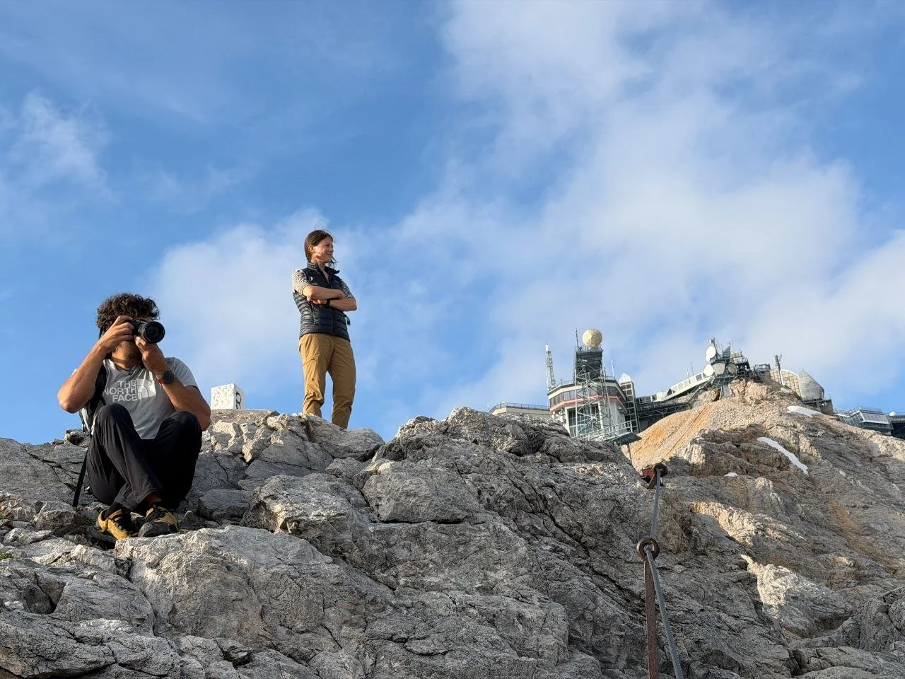 Two hikers on a rocky mountain trail, one woman standing with arms crossed and another man sitting and taking a photo, with an observation station or communication building with antennas in the background under a blue sky with clouds.