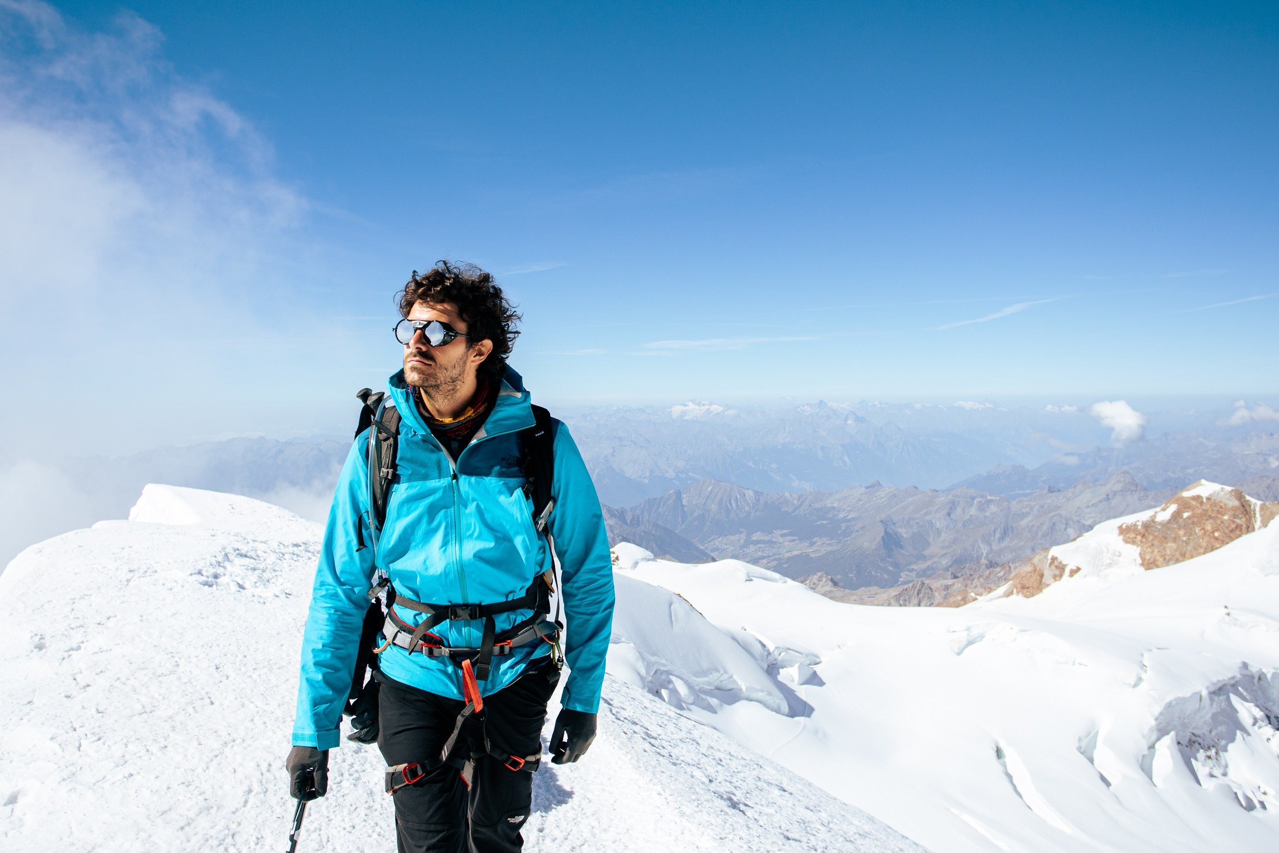A man wearing sunglasses, a blue jacket, and a backpack hiking on snowy mountain terrain with mountains in the background and a clear blue sky.