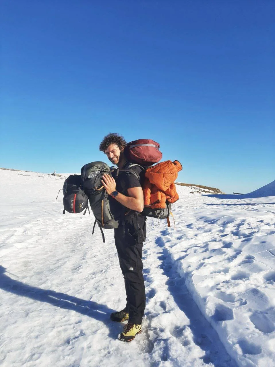 A man with curly hair smiling and carrying a large backpack with outdoor gear, standing on snow with a clear blue sky in the background.