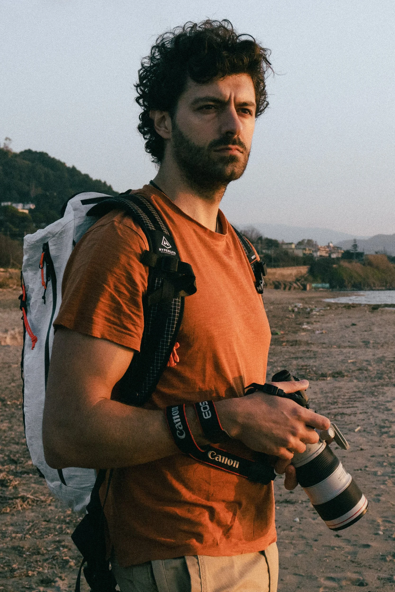 A man with curly dark hair and a beard holding a camera lens in his hands, wearing an orange shirt and a backpack, standing outdoors near the coast during sunset.