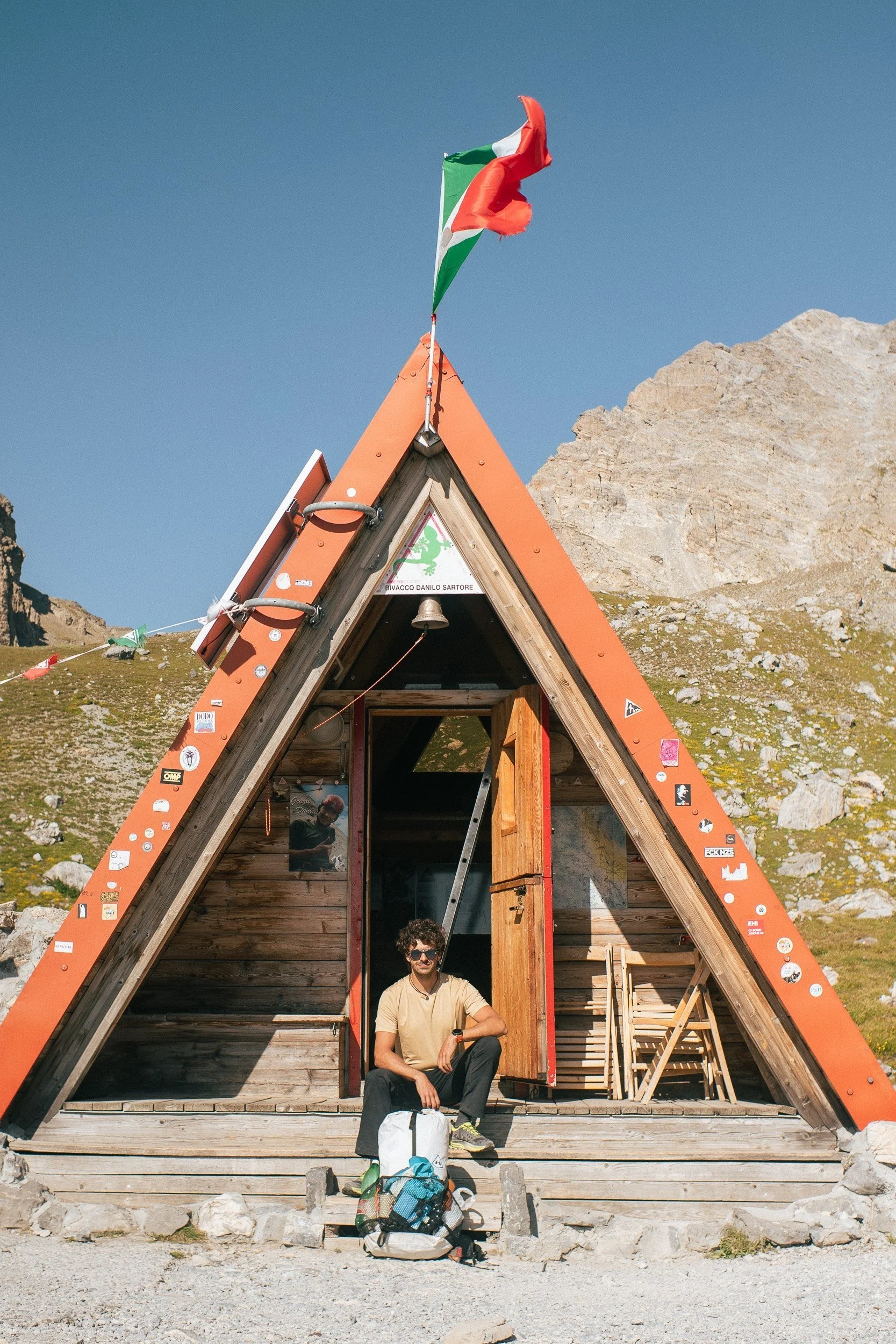 Man sitting in front of an A-frame wooden structure with a triangular roof, covered in stickers and a flag on top, situated in a mountainous landscape with rocky terrain and clear blue sky.