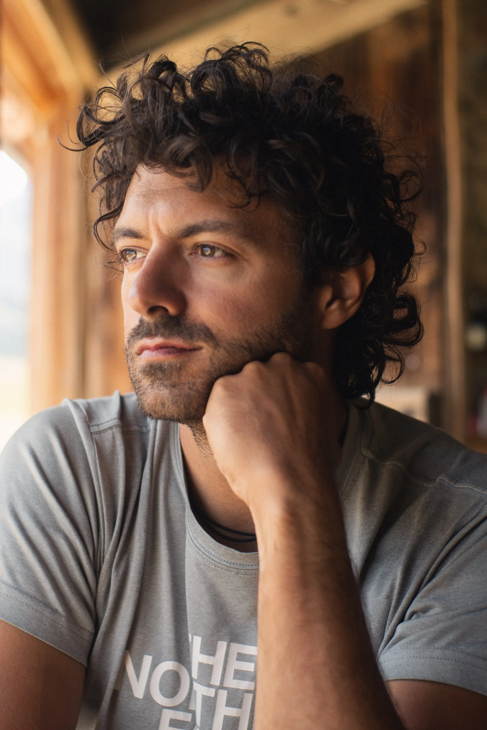 Close-up of a man with curly dark hair and facial hair, resting his chin on his hand, looking out a window with a thoughtful expression, wearing a gray t-shirt with partly visible text.