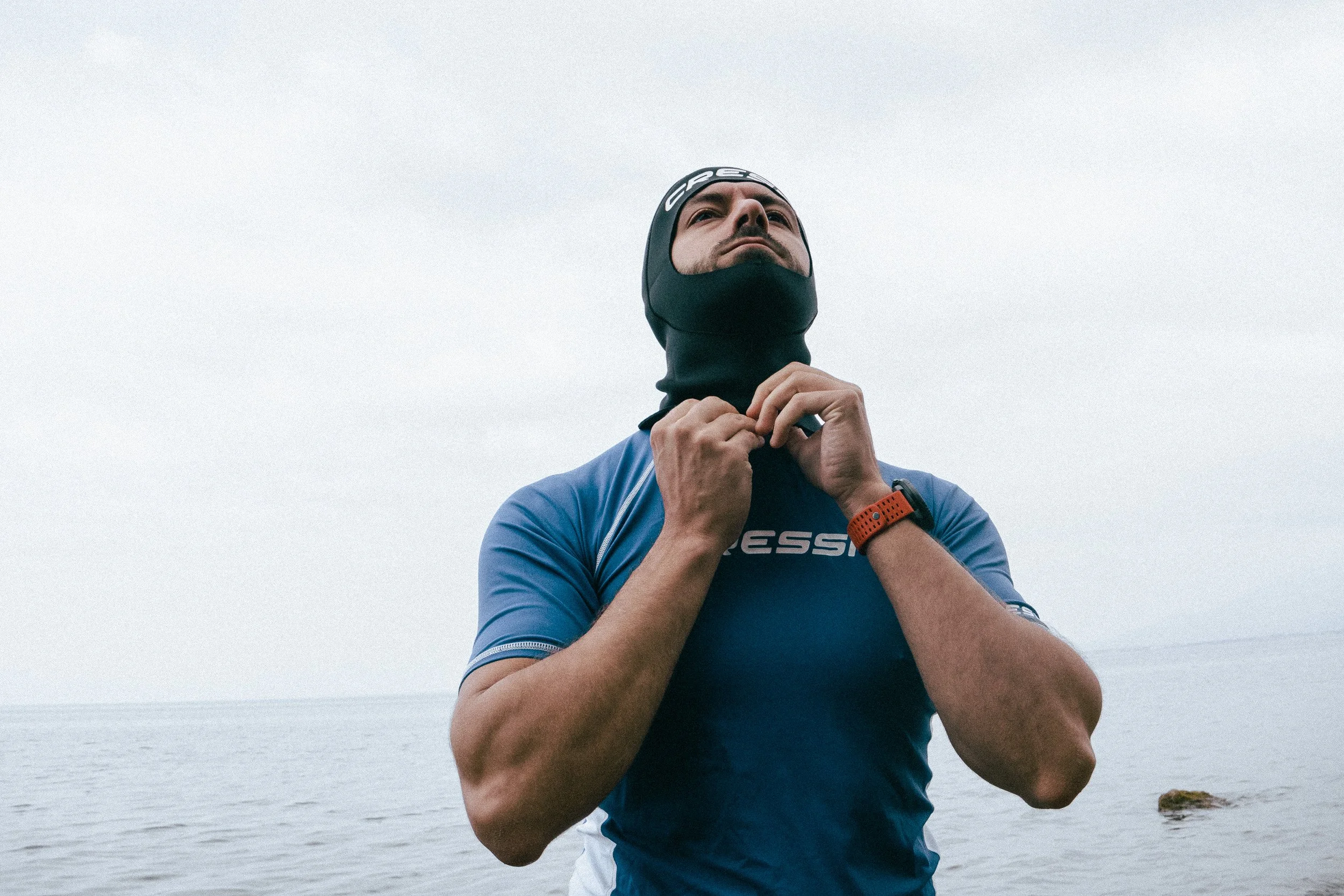 A man outdoors adjusting a neck gaiter or face covering, wearing athletic clothing and a watch, with a body of water and a cloudy sky in the background.