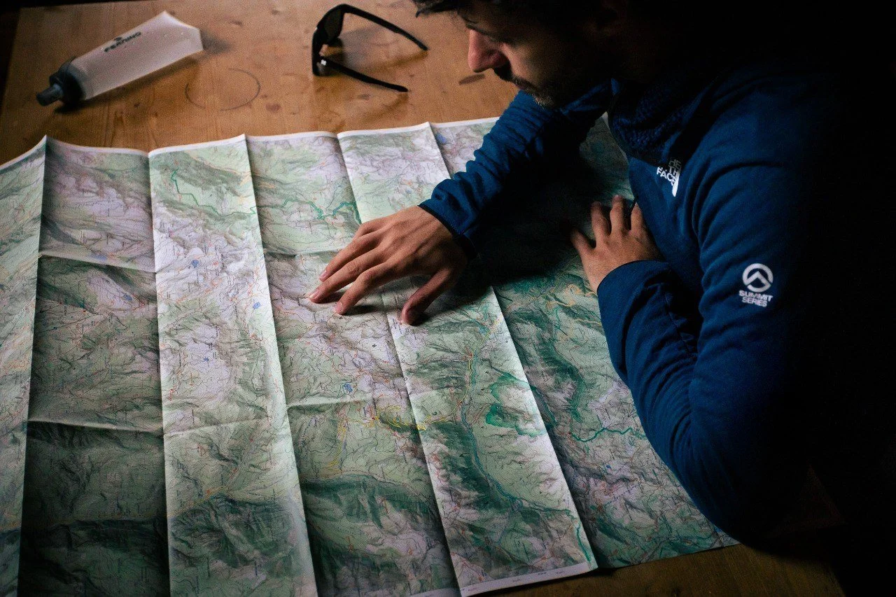 Person in a blue jacket looking at a folded topographical map on a wooden surface, with a pair of glasses and a tube of cream nearby.