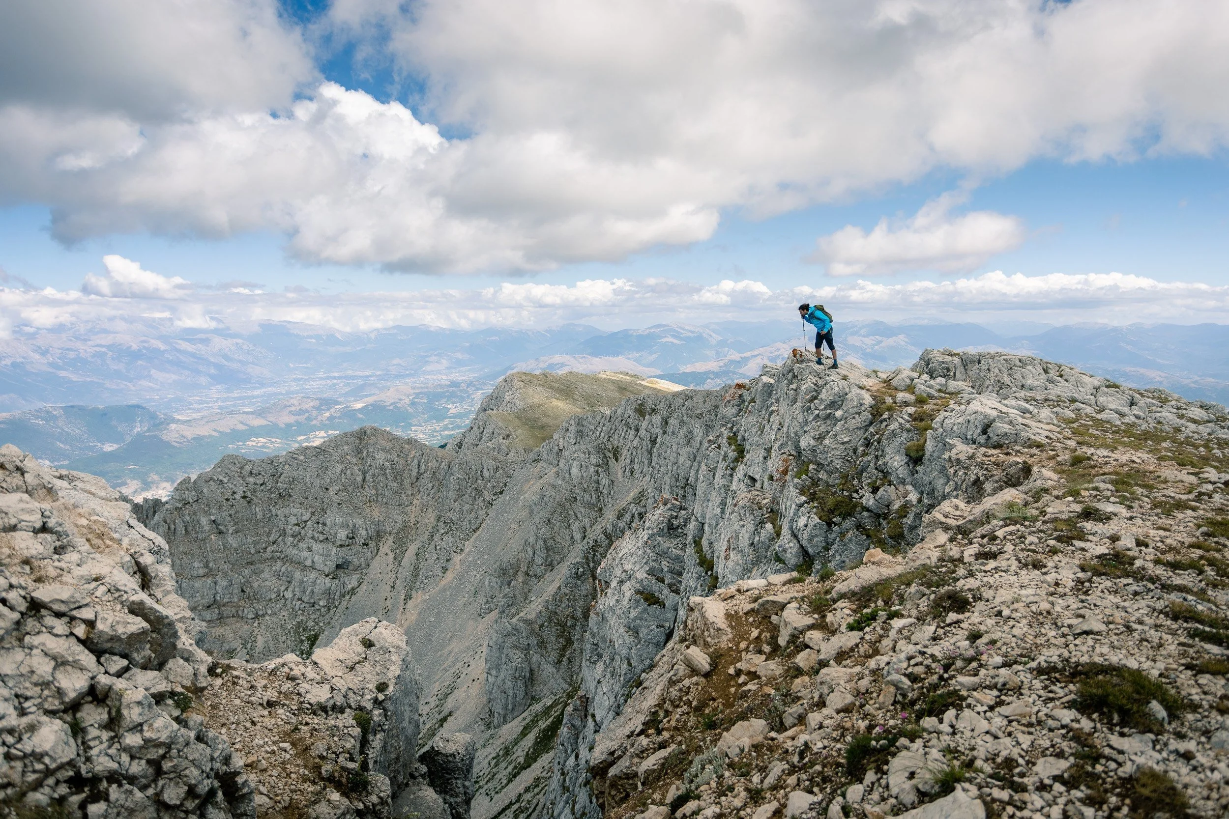 Hiker wearing a blue jacket and backpack standing on rocky mountain ridge with expansive view of valleys and distant mountains under cloudy sky.