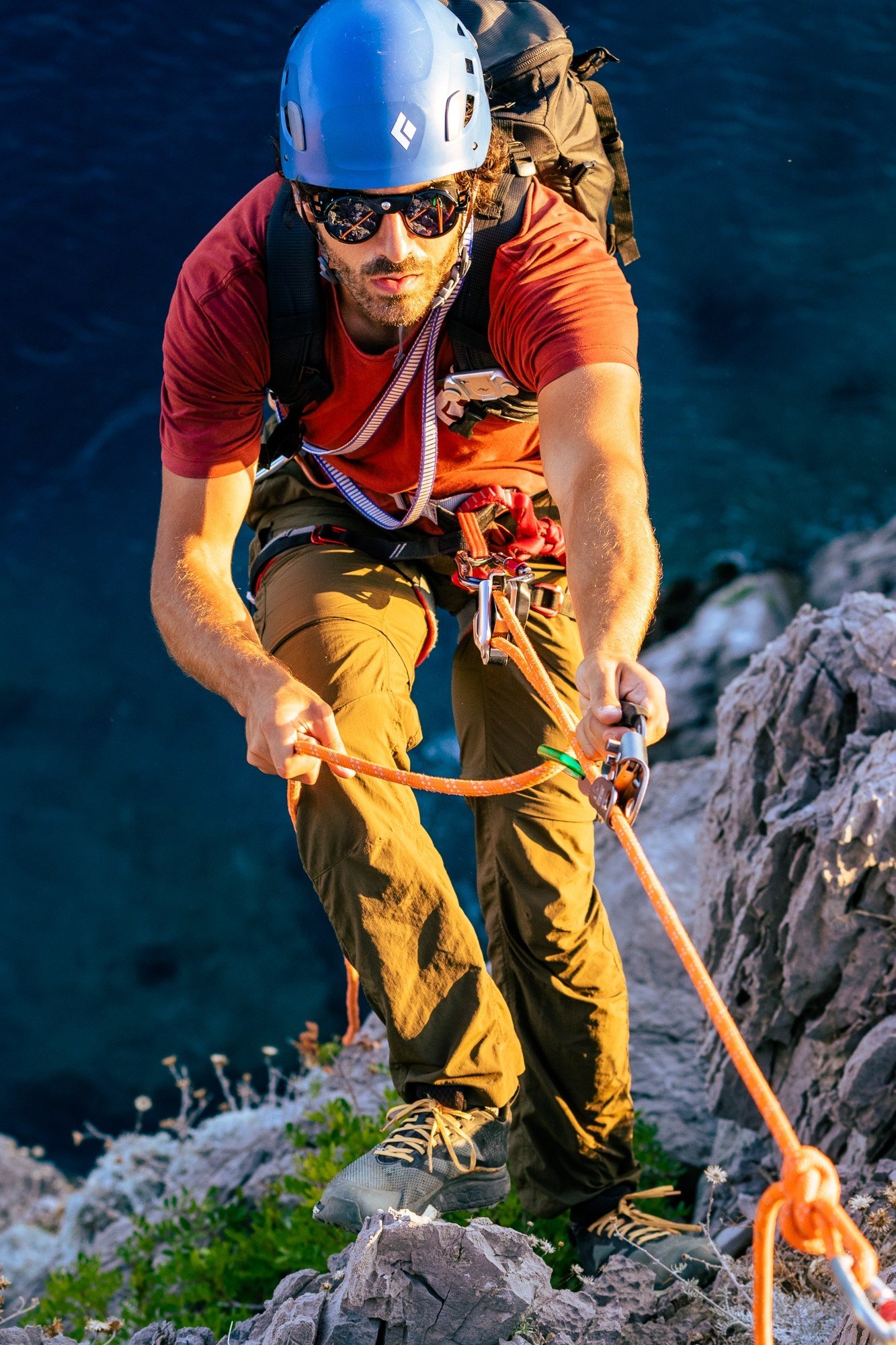 A man wearing a helmet, sunglasses, and climbing gear rappelling down a rocky cliff face with the ocean in the background.
