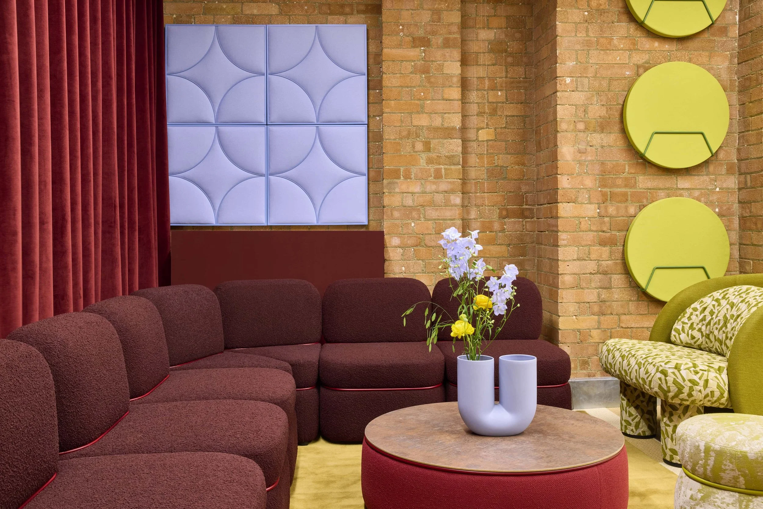 A bold lounge space, a modular burgundy sofa sits in front of a deep red velvet curtain. A olive rug is below and by the side of the central coffee table there is an armchair that is upholstered in a graphic green pattern.