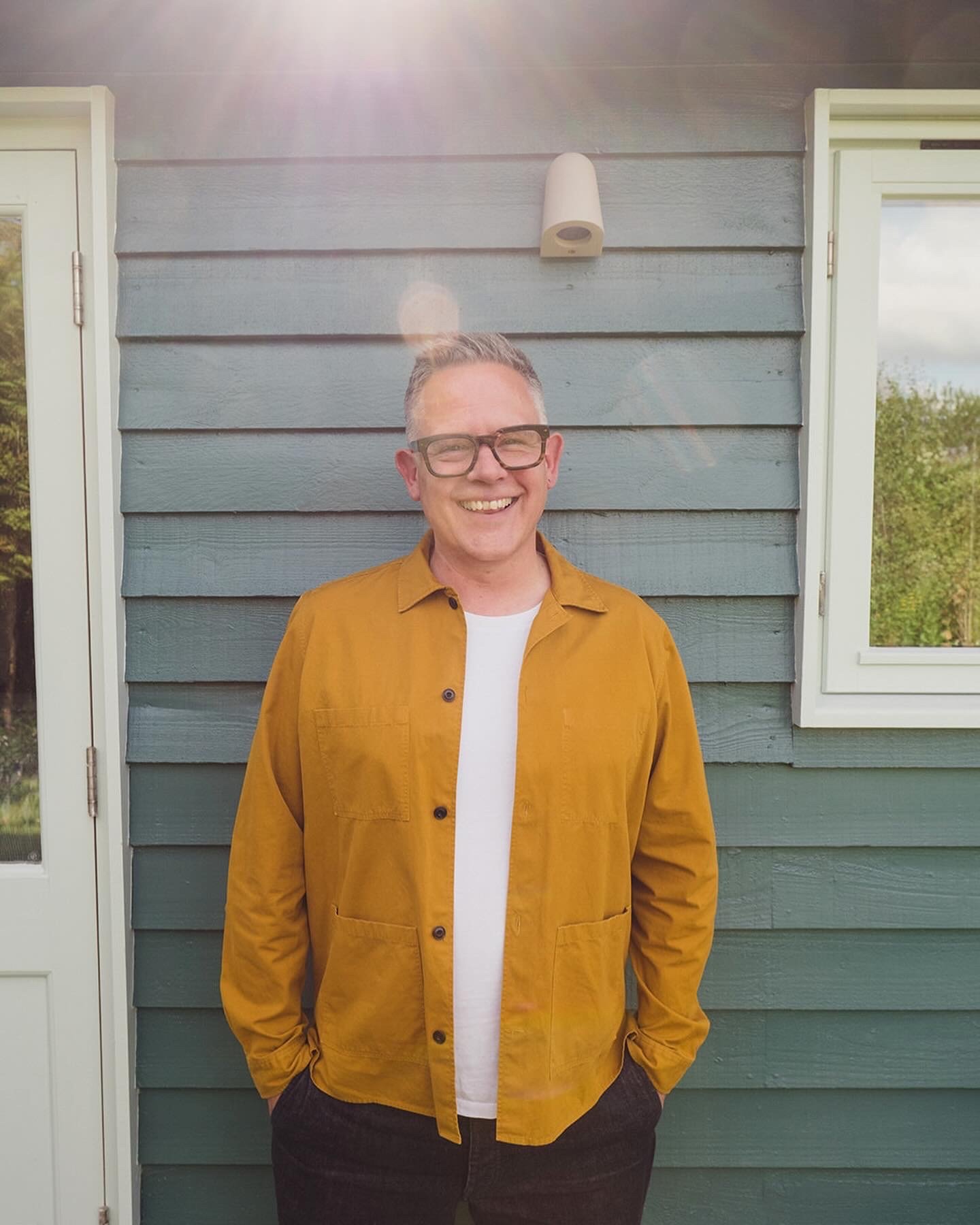 Image of a smiling white man with glasses. He wears a white t-shirt with a mustard shirt over the top and stands in front of a garden room