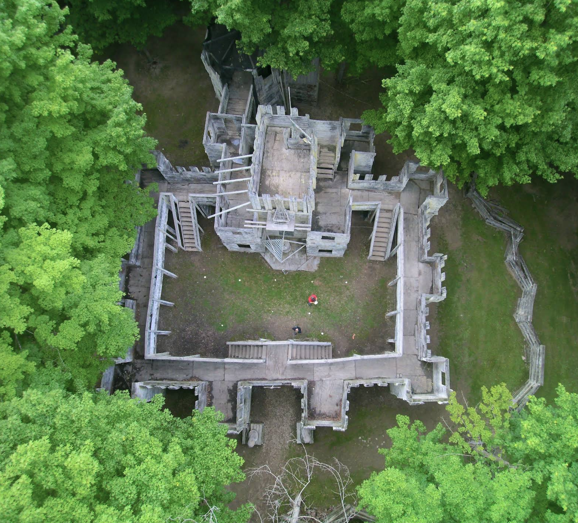 Aerial view of an abandoned wooden castle surrounded by green trees, with two people visible at the bottom.