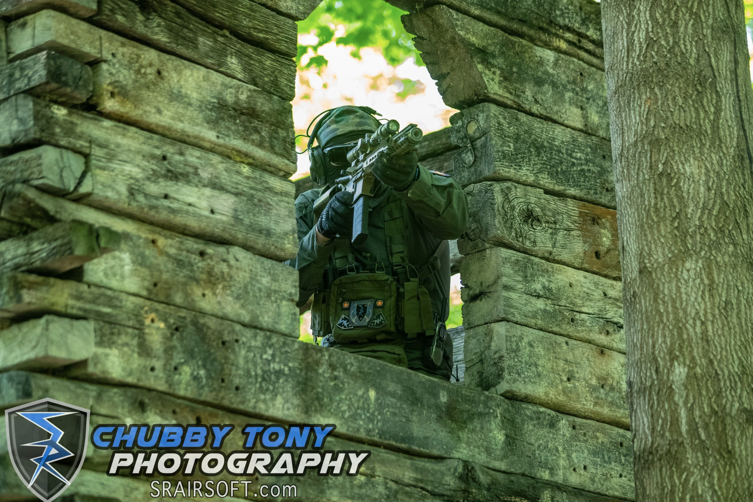 A person dressed in tactical gear aiming a rifle through a wooden structure, with a background of green foliage.
