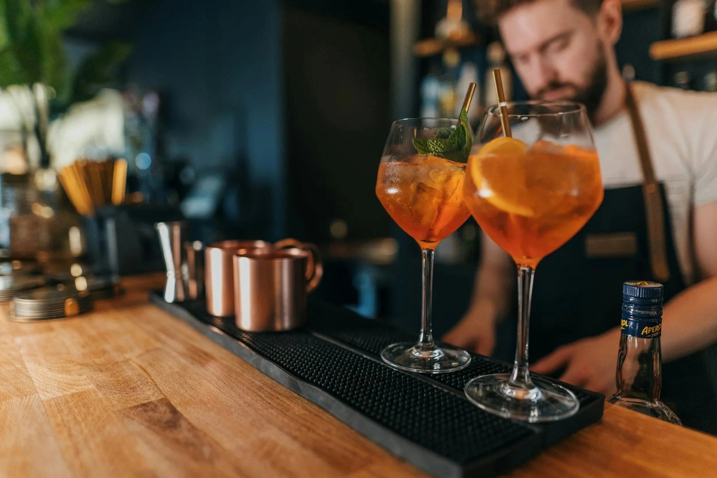 Two orange-colored mocktails garnished with lemon slices and mint leaves on a bar counter, with a bartender in the background.