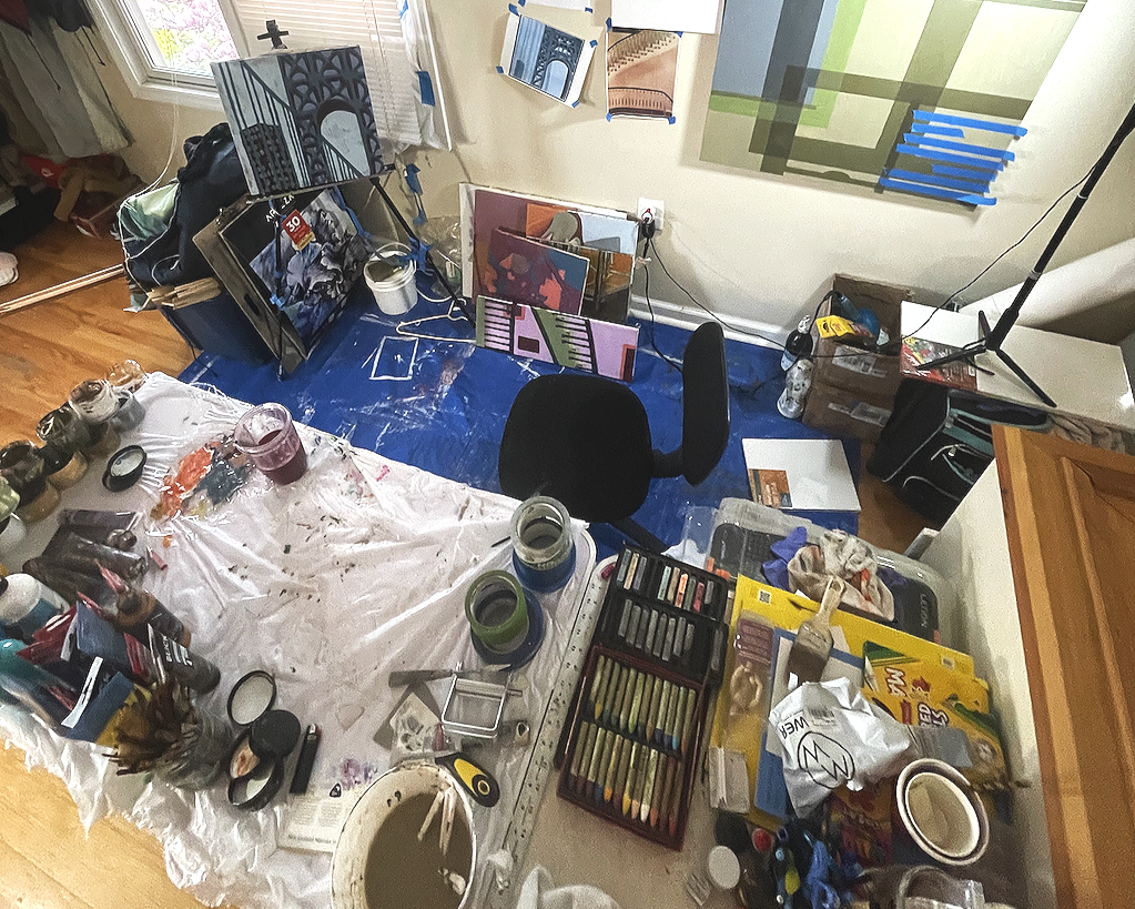 Artist's workspace with paints, brushes, and canvases, cluttered with art supplies, tables, chairs, and artworks leaning against walls.