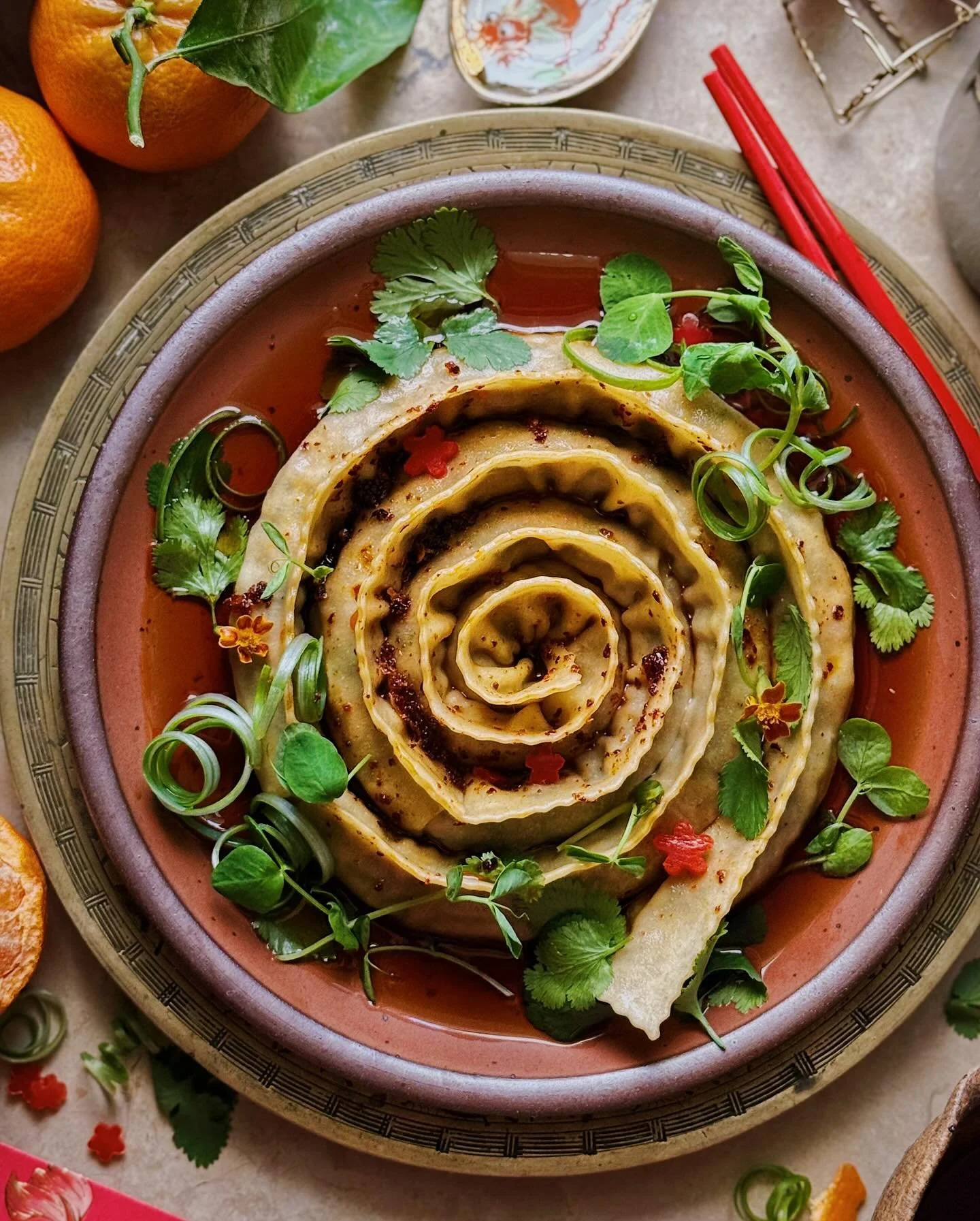 Jumbo spiral dumplings filled with pork, seasonal greens, and shiitake mushrooms; resting in a sauce of soy, citrus, and @california.wines sparkling wine; topped with a drizzle of chili oil and some pea shoots, cilantro, scallions, and tiny flowers c