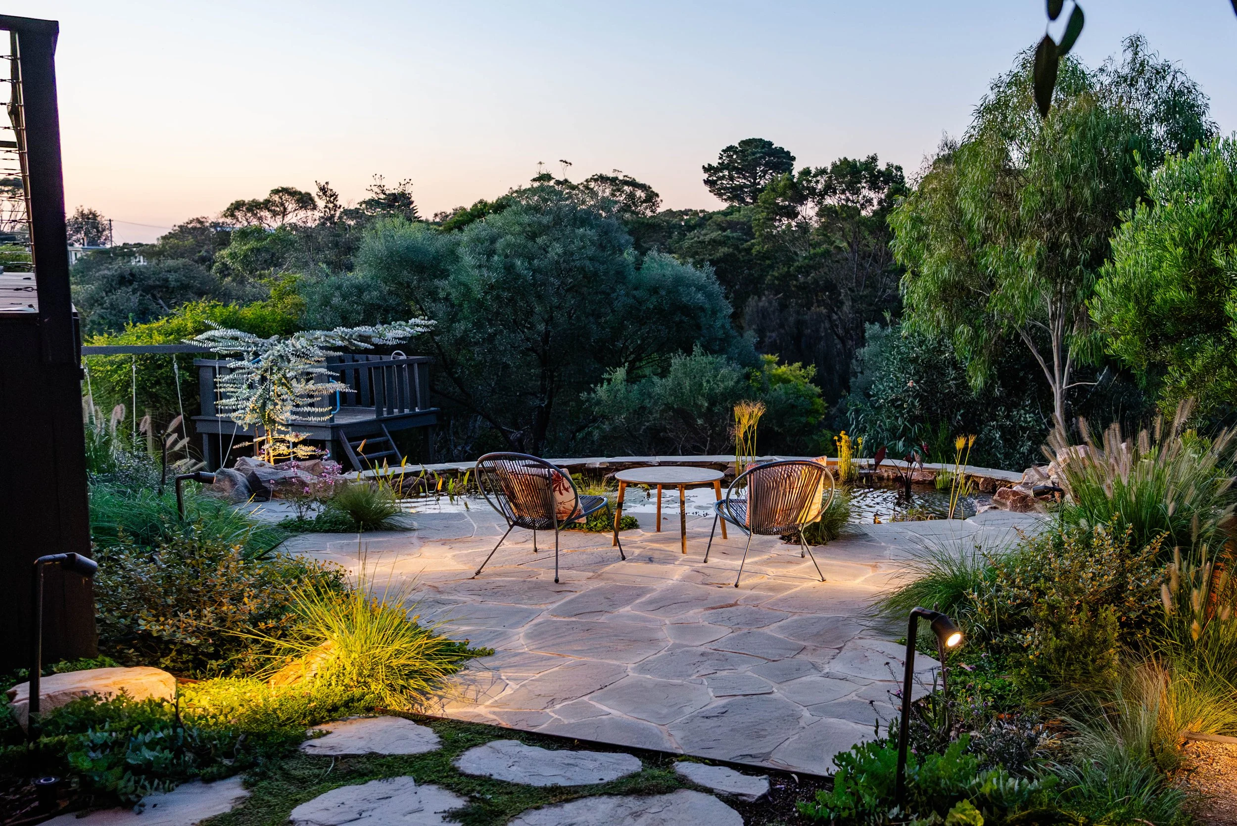 Mornington Peninsula, Mount Martha backyard paved patio overlooking a natural pond with two lounge chairs and a round table illuminated by outdoor lights, surrounded by green plants and trees at dusk designed by sheoak design.