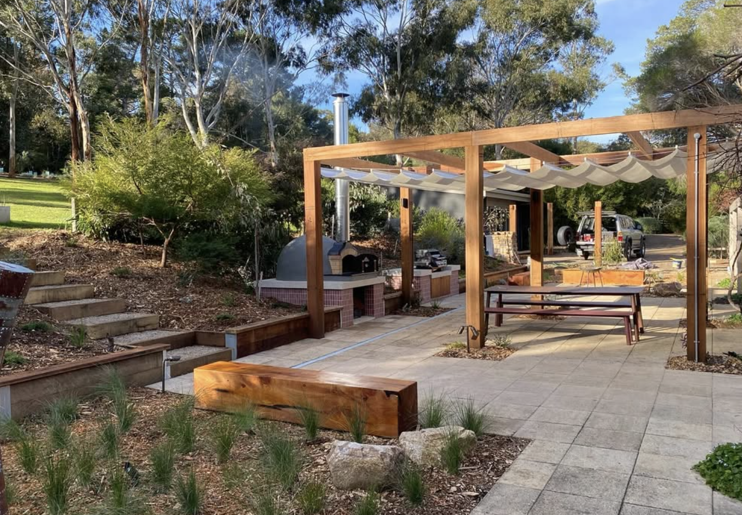 Mornington Peninsula, Shoreham, Backyard patio area with a wood pergola, outdoor pizza oven, a picnic table, and a garden, with a driveway and a black SUV in the background, surrounded by trees and greenery designed by Sheoak Design.