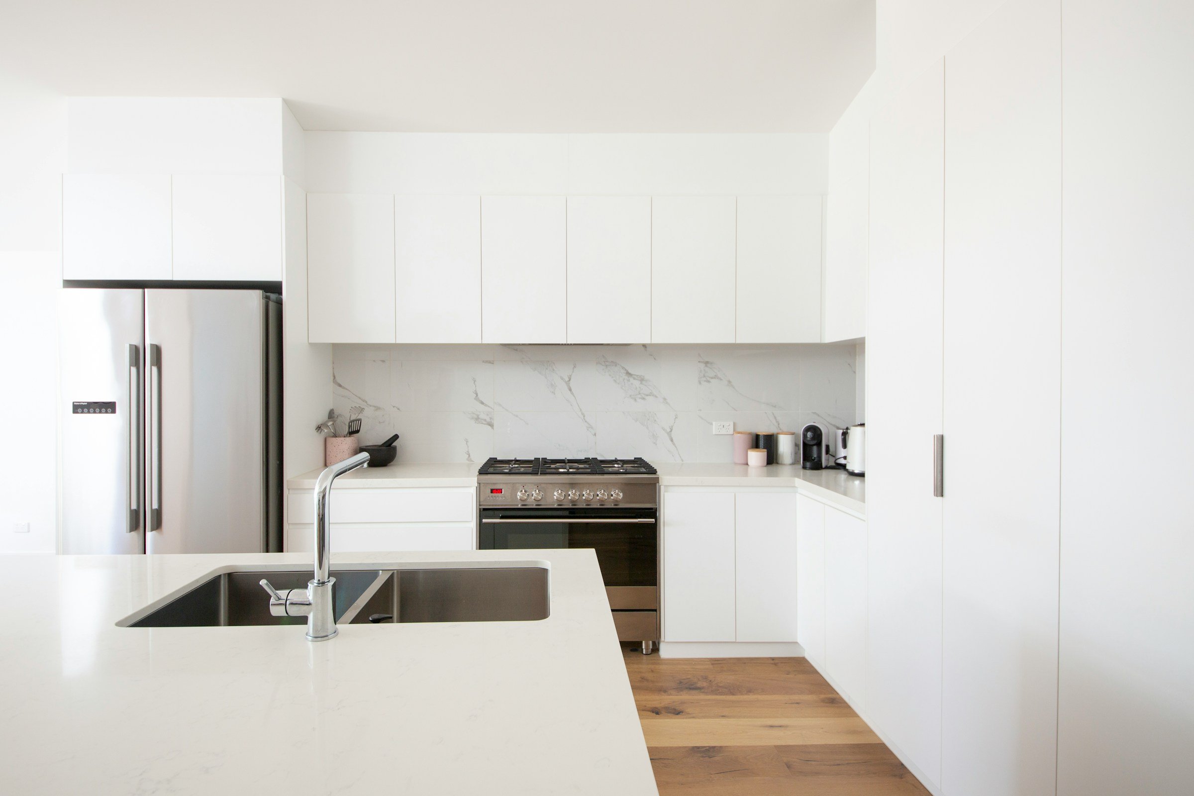 Modern kitchen with white cabinets, a stainless steel refrigerator, a stove, and a marble backsplash. An island with a white countertop and an undermount sink with a chrome faucet is in the foreground. Small kitchen appliances and utensils are on the counters.