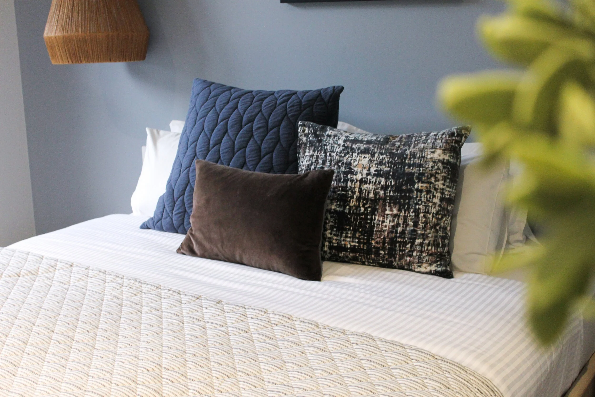 A neatly made bed with striped bedding and four decorative pillows in blue, black and white, and brown, positioned against a light blue wall, with a partially visible wooden headboard and a green plant in the foreground.