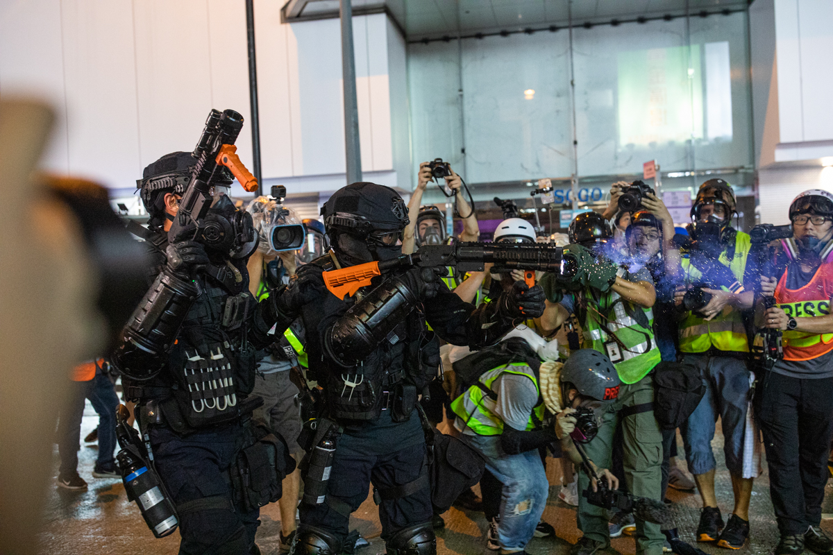 Cops shooting rubber bullets while journalists look on with genuine facial expressions.