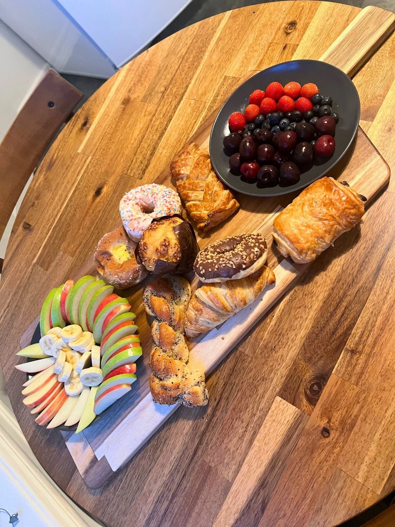 High-angle shot of a rectangular wooden serving board on a round wooden table, arranged with assorted pastries.