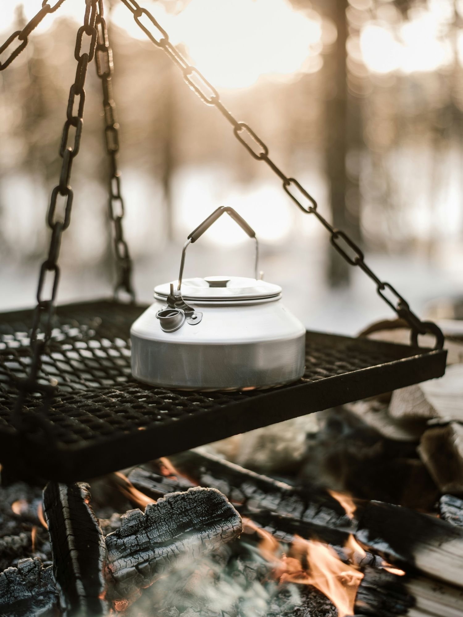 A close-up shot of a classic brushed aluminum camping kettle sitting on a black metal mesh grill grate. The grate is suspended by sturdy black chains directly over a crackling campfire with burning logs and orange flames.