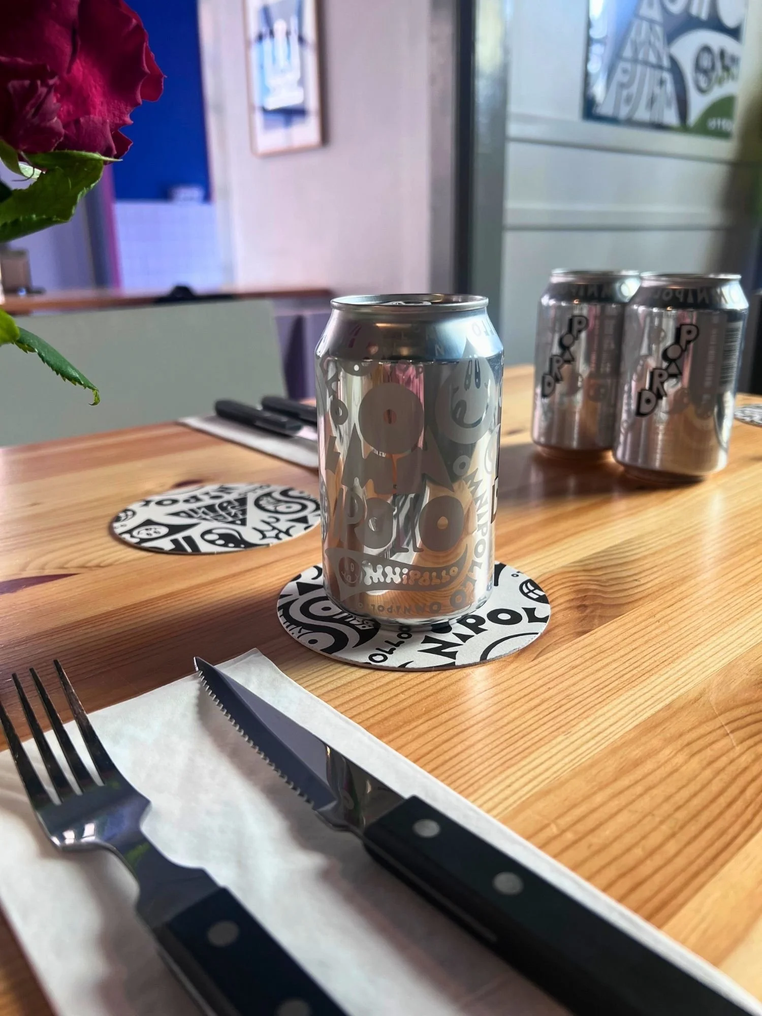 A metallic silver beer can on a round, black-and-white patterned Omnipollo coaster on a light wooden table. In the foreground, a fork and a black-handled steak knife on a white napkin.