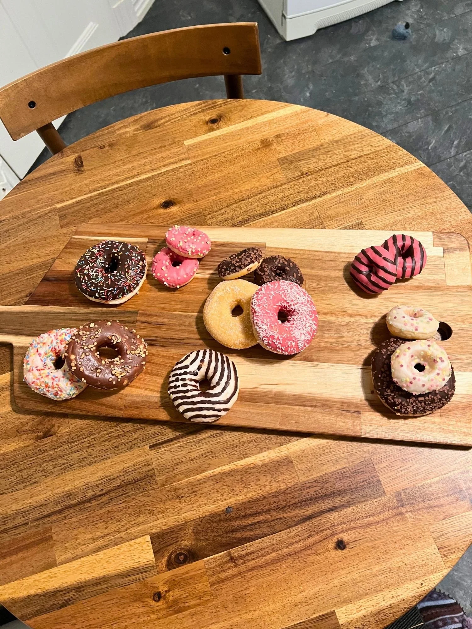 High-angle shot of a rectangular wooden serving board on a round wooden table, displayed with an assortment of glazed, frosted, and sprinkled donuts and mini donuts.