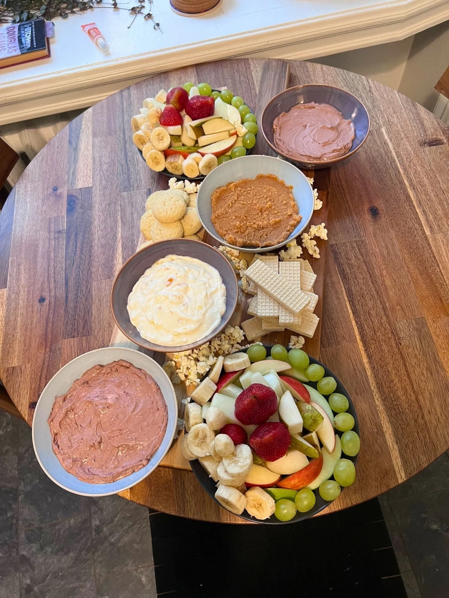 High-angle view of a wooden snack board featuring two fruit platters, four bowls of dessert dips, round cookies, stacked wafer biscuits, and scattered popcorn.