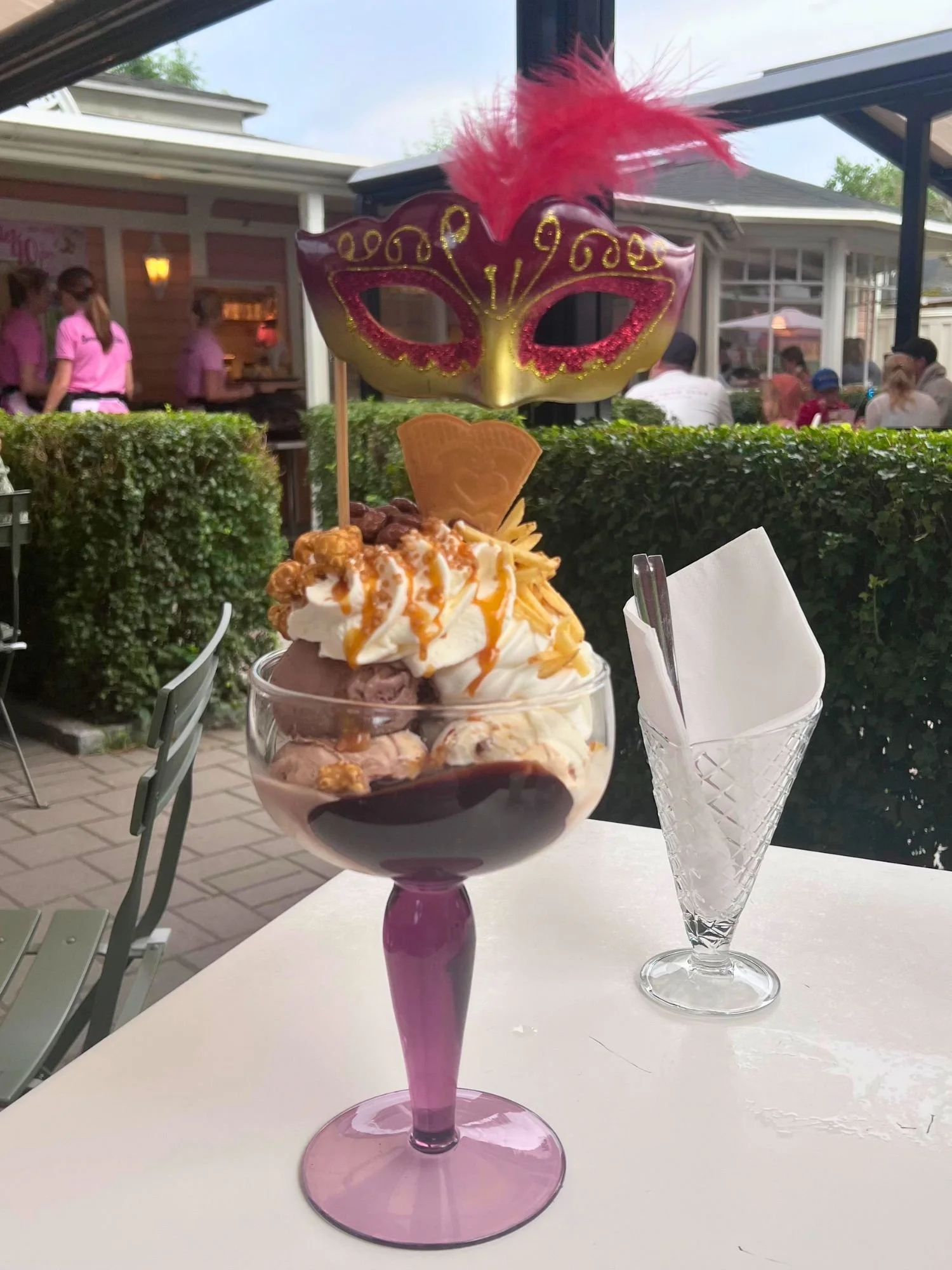 An elaborate ice cream sundae in a purple-stemmed glass, topped with whipped cream, caramel, and a decorative red masquerade mask.