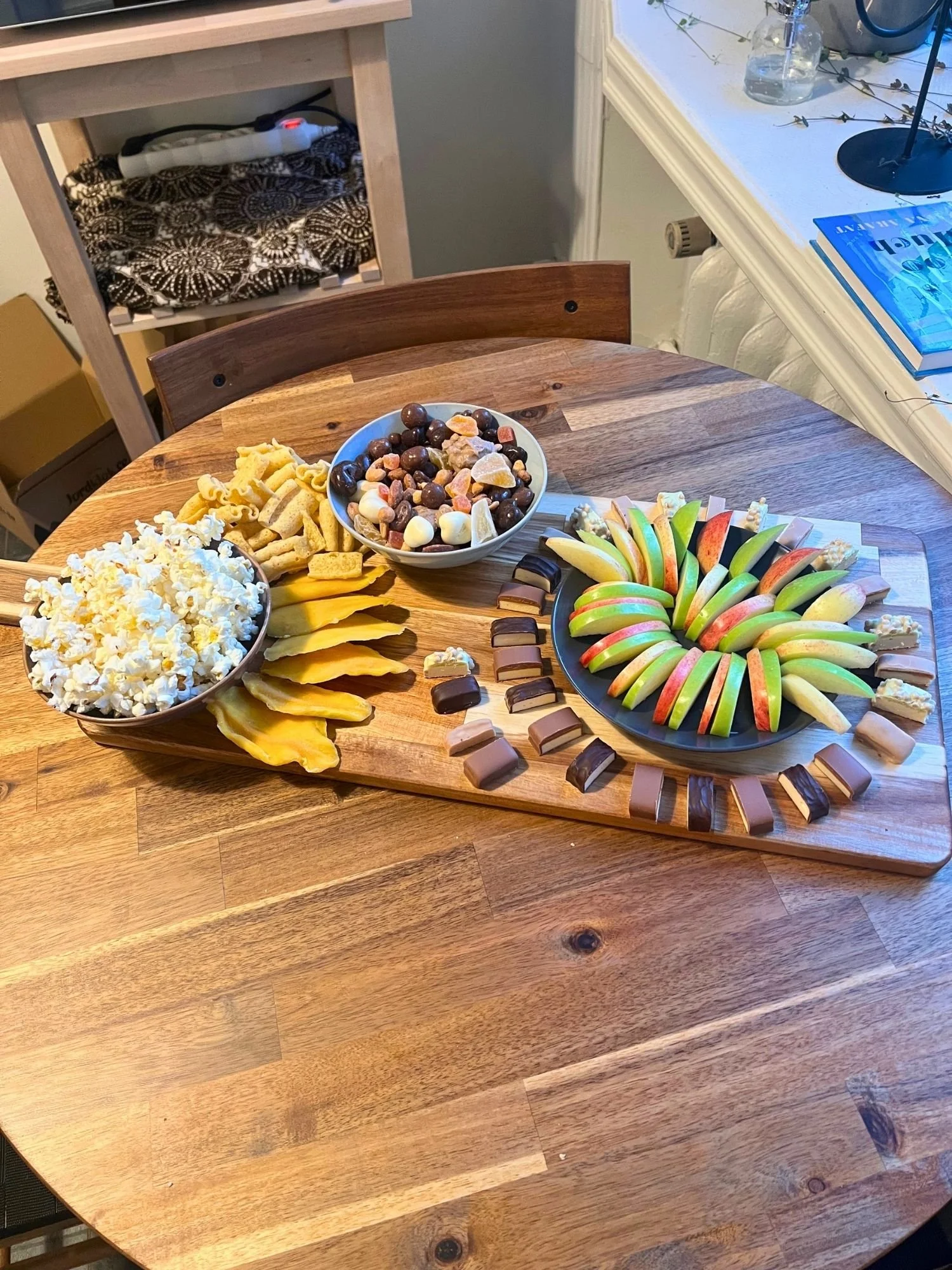 A wooden table set with a snack board featuring a bowl of popcorn, trail mix, dried fruit slices, a ring of sliced apples, and small protein bar bites.