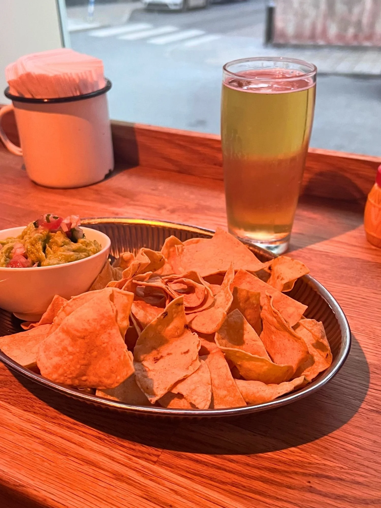 A round metal tray filled with crisp tortilla chips on a wooden counter next to a small white bowl of guacamole topped with diced tomatoes. Behind the chips stands a pint glass of clear, light golden beer and an enamel mug holding napkins.