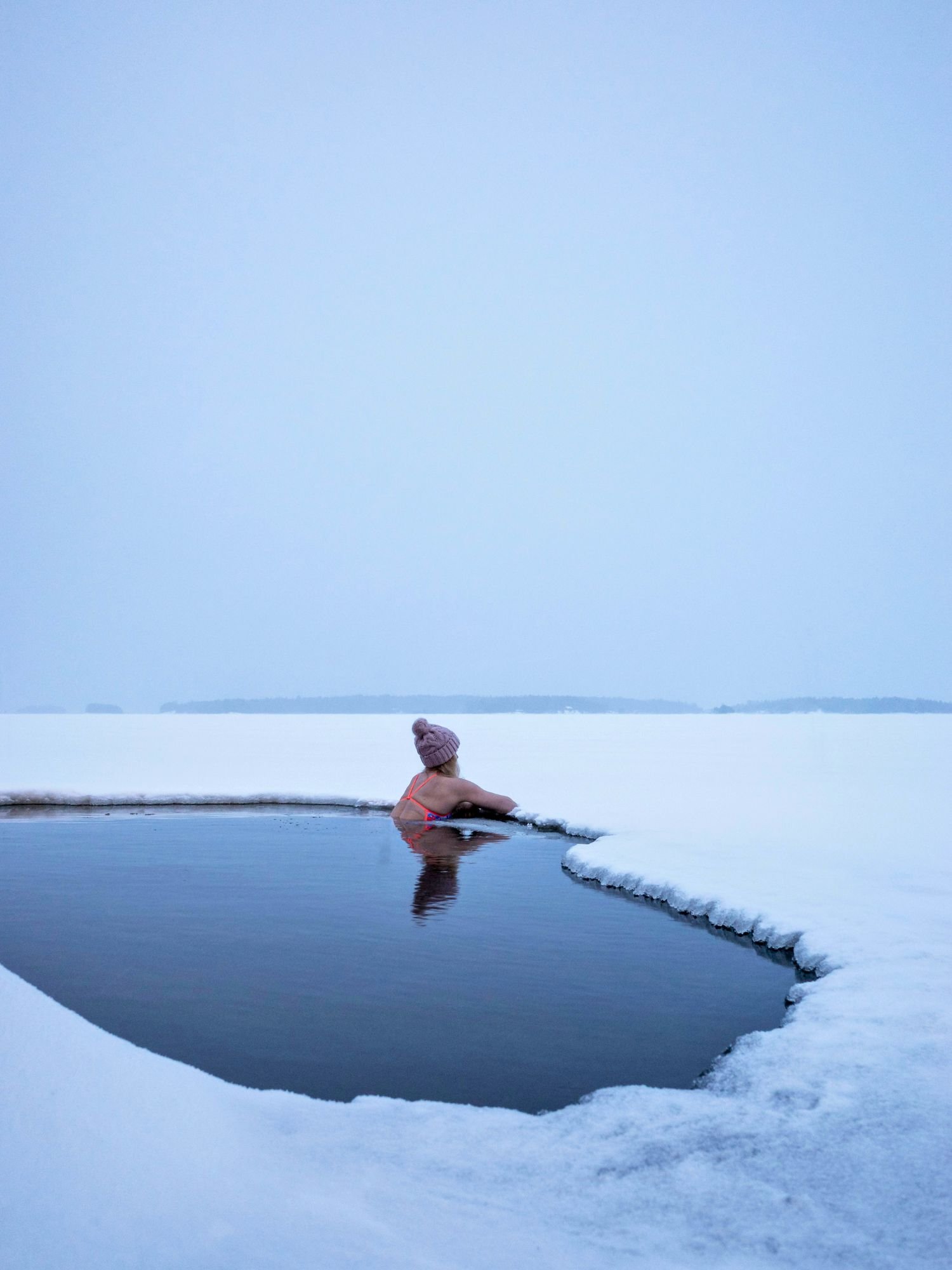 Woman in a winter hat sits in a hole cut into a frozen lake, surrounded by snow and ice during an outdoor ice swimming experience.
