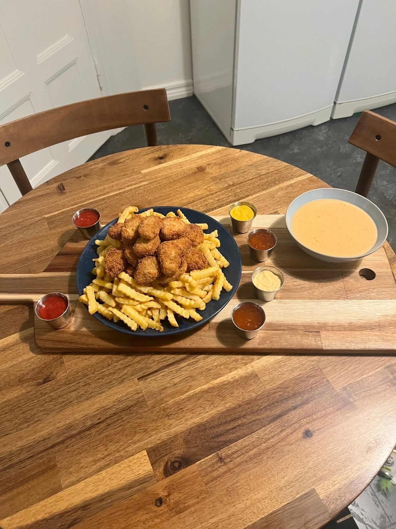 High-angle shot of a long wooden serving board on a round wooden table, set with a blue plate overflowing with crinkle-cut fries and breaded nuggets, accompanied by small dipping sauce cups and a bowl of cheese sauce.