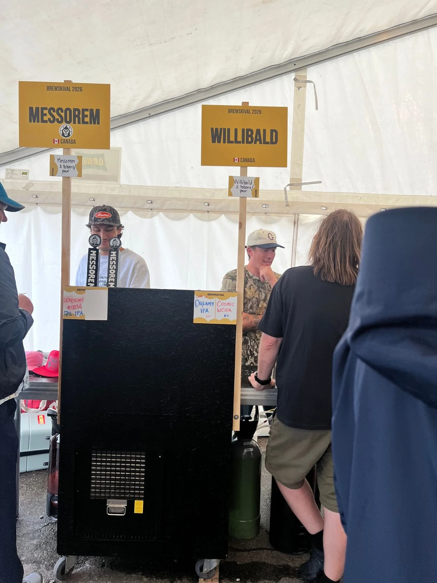 A beer festival booth inside a white tent with yellow overhead signs for Messorem - Canada and Willibald - Canada. Behind the counter, staff members interact with festival attendees, with tap handles visible.
