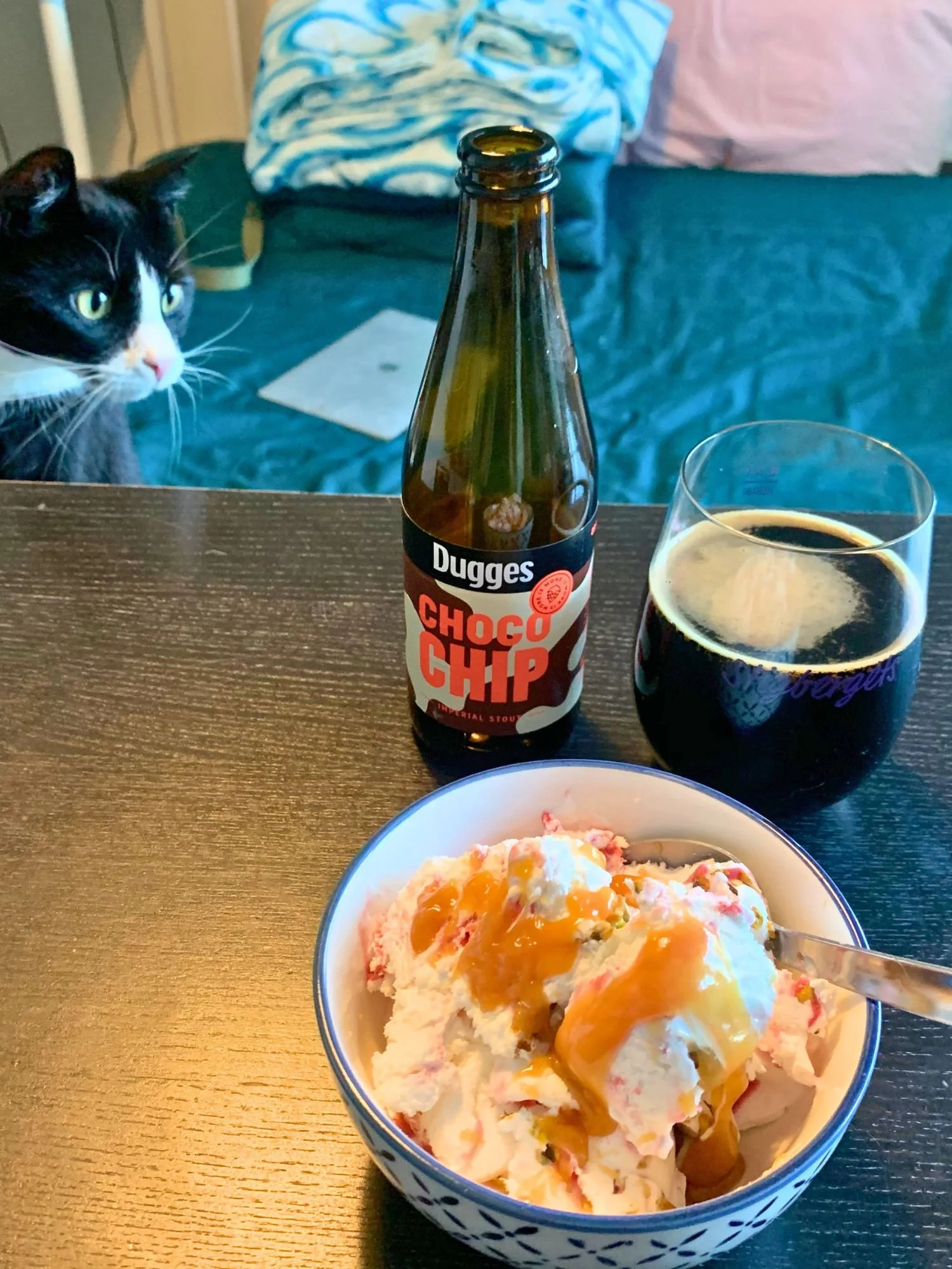 A dark wood table featuring a bowl of white and pink ice cream topped with caramel sauce, an open bottle of Dugges Choco Chip Imperial Stout, and a stemless glass of dark beer.