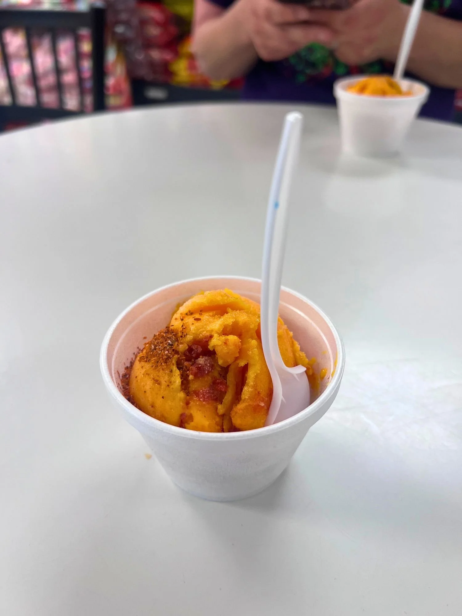 A white styrofoam cup filled with vibrant orange mango ice cream, sprinkled with chili powder seasoning and served with a white plastic spoon on a white table.