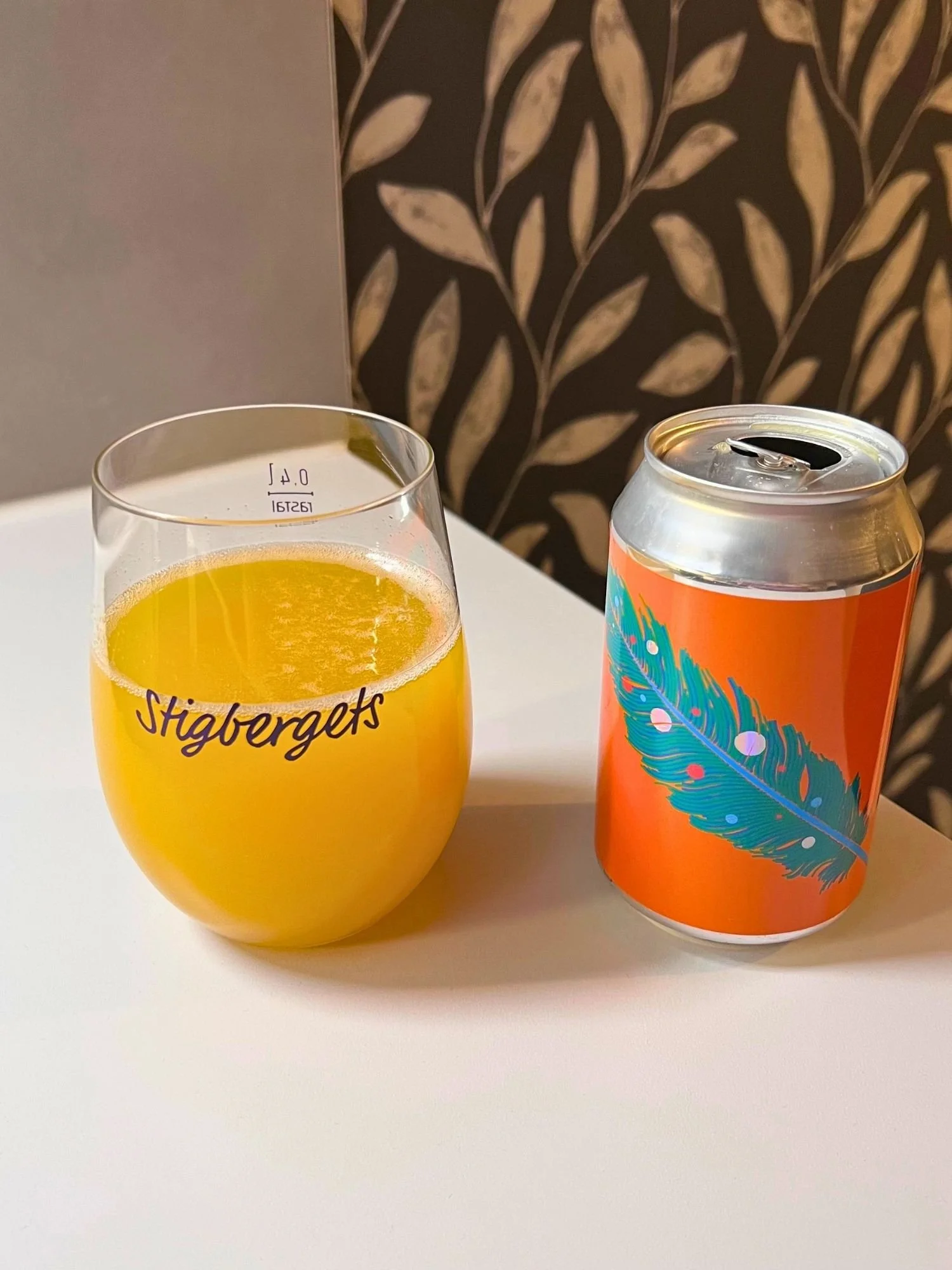 An opened beer can with a bright orange label featuring a teal feather standing on a white surface next to a stemless glass filled with thick, bright yellow sour beer. The glass is printed with the cursive text "Stigbergets".