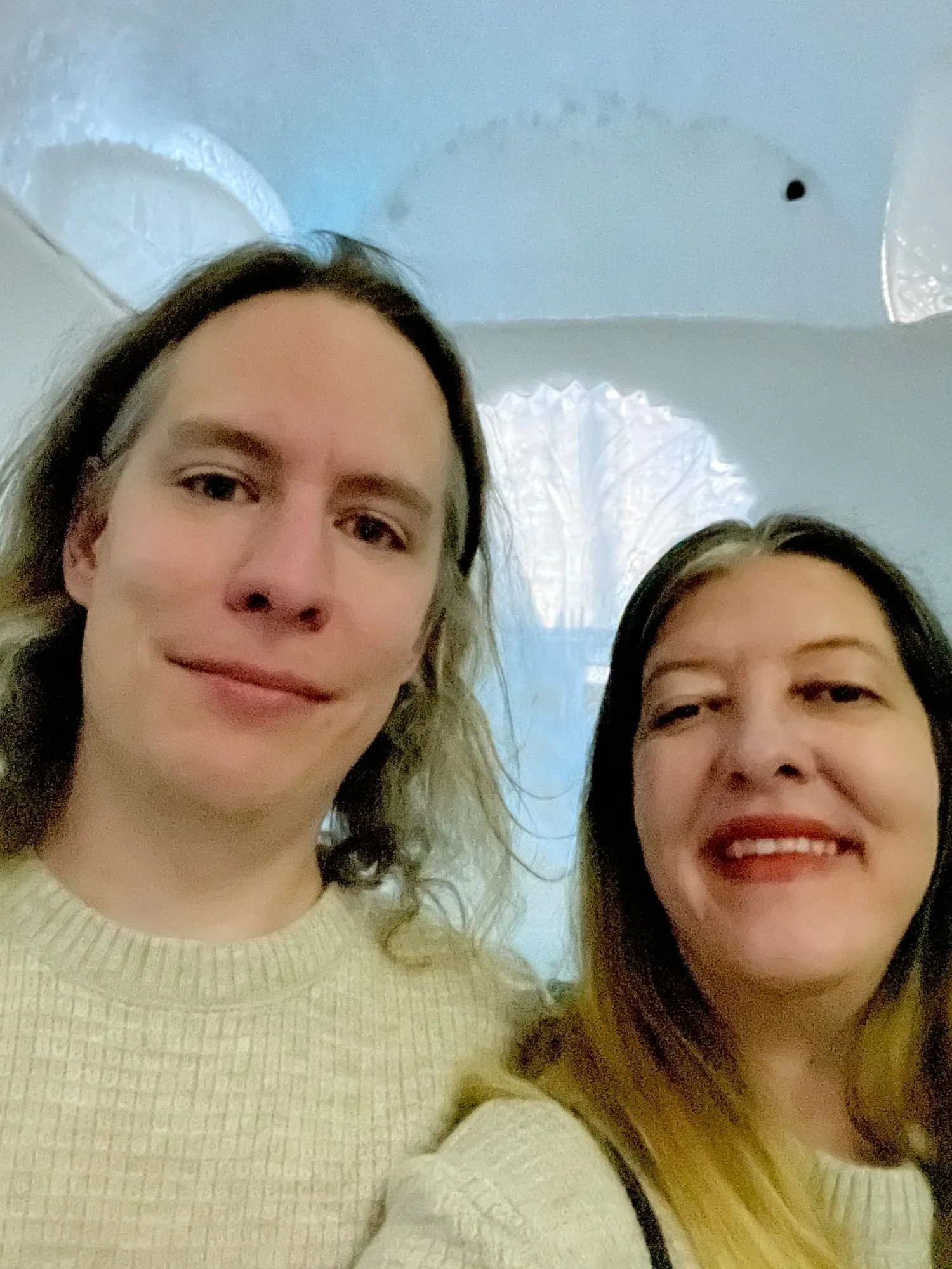Two people smiling for a selfie inside an ice hotel room, with carved ice walls and soft blue lighting in the background.