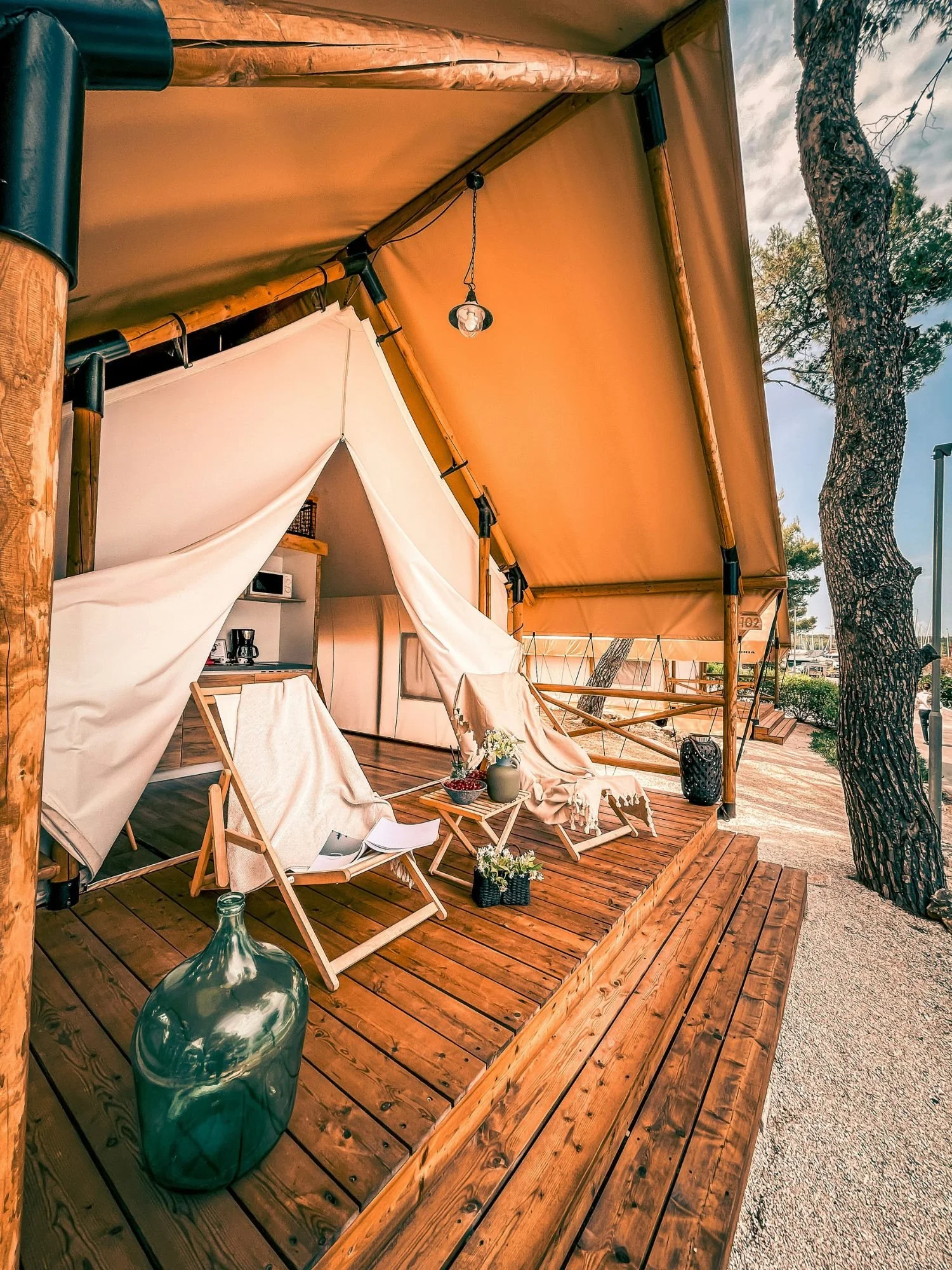 A large luxury glamping tent built with thick wooden poles and a tan canvas roof canopy sits on a raised wooden deck with wide steps.