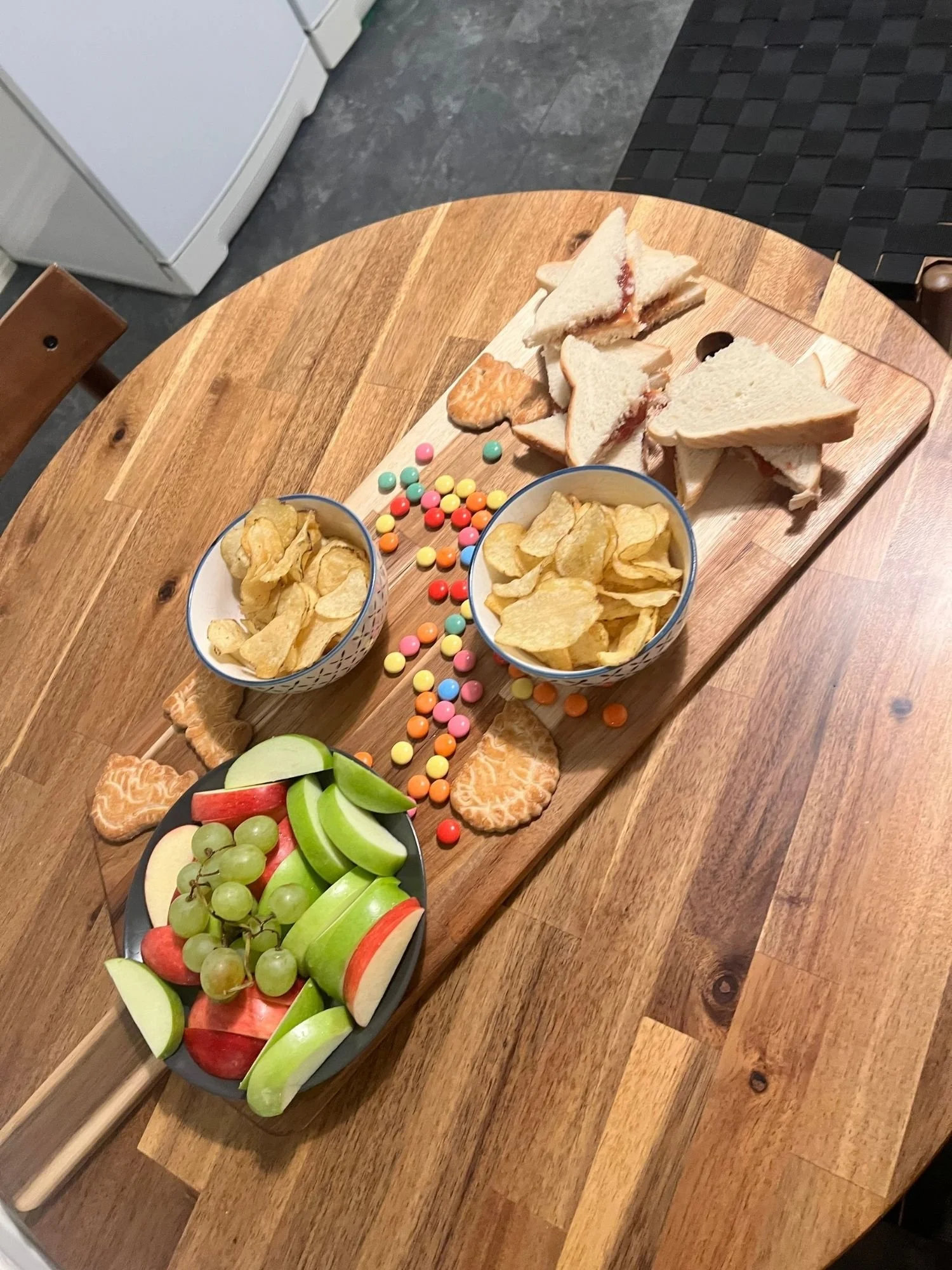 High-angle shot of a long wooden serving board on a round wooden table, set with a child-friendly spread featuring sandwich triangles.